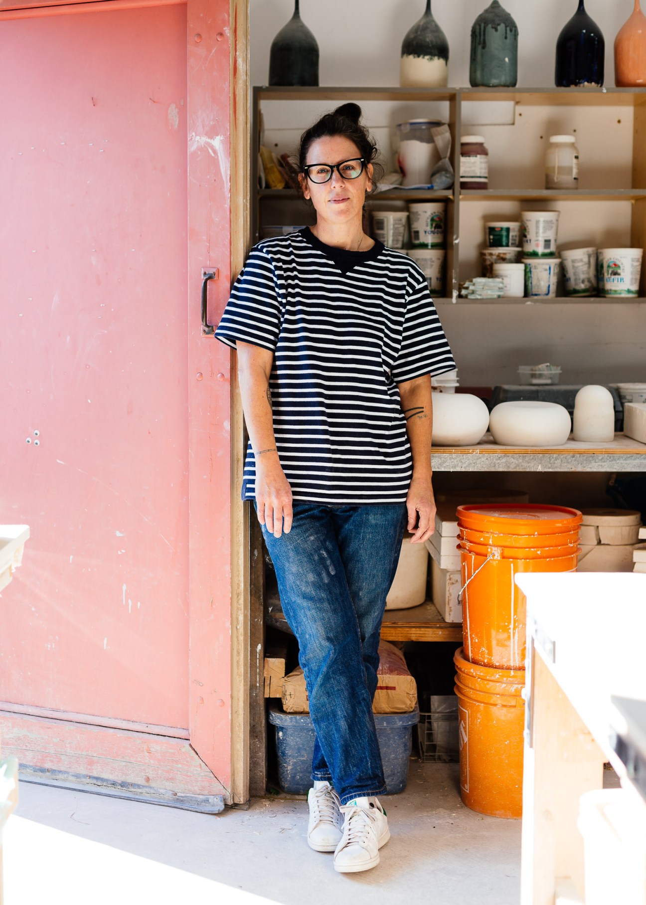 Woman in striped shirt standing in front of wooden shelving with ceramics and home goods in a studio.