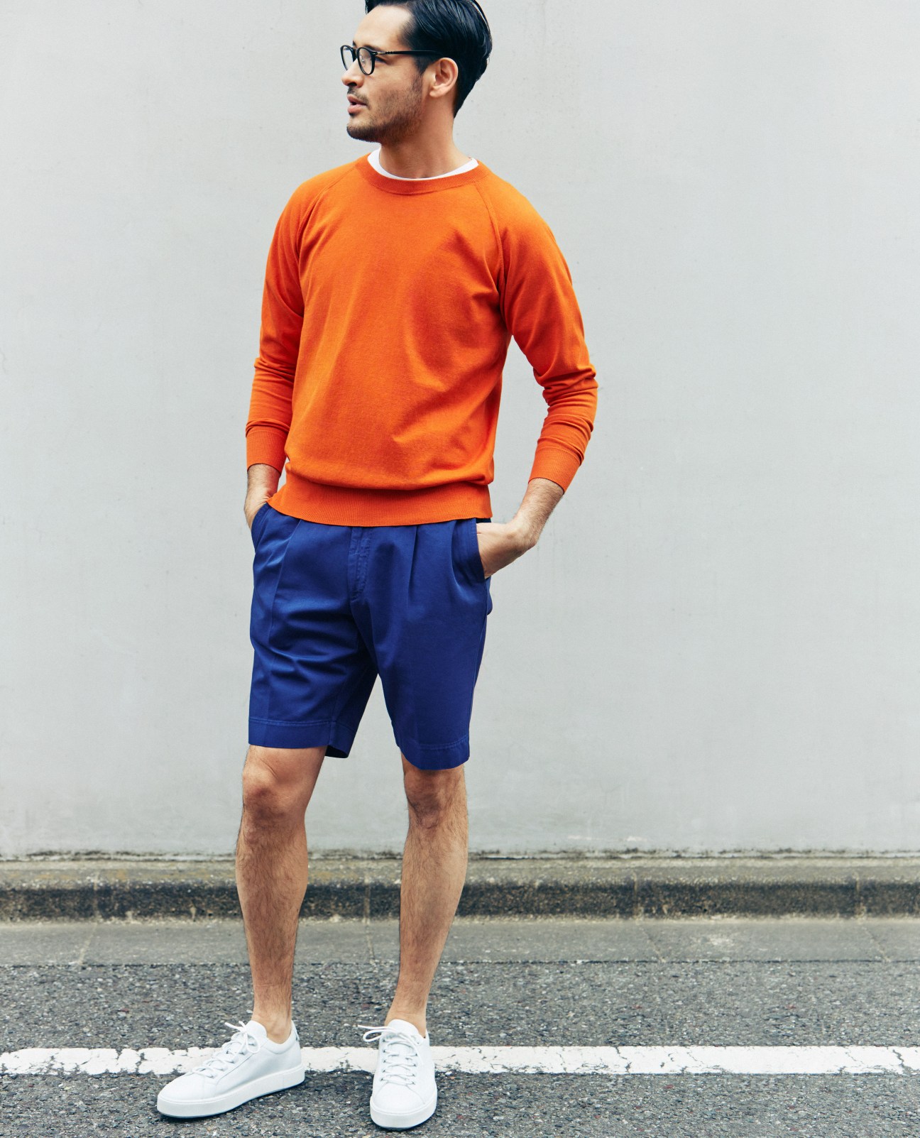 Man wearing orange crewneck sweater, navy shorts, white trainers, and tortoiseshell glasses standing outdoors.