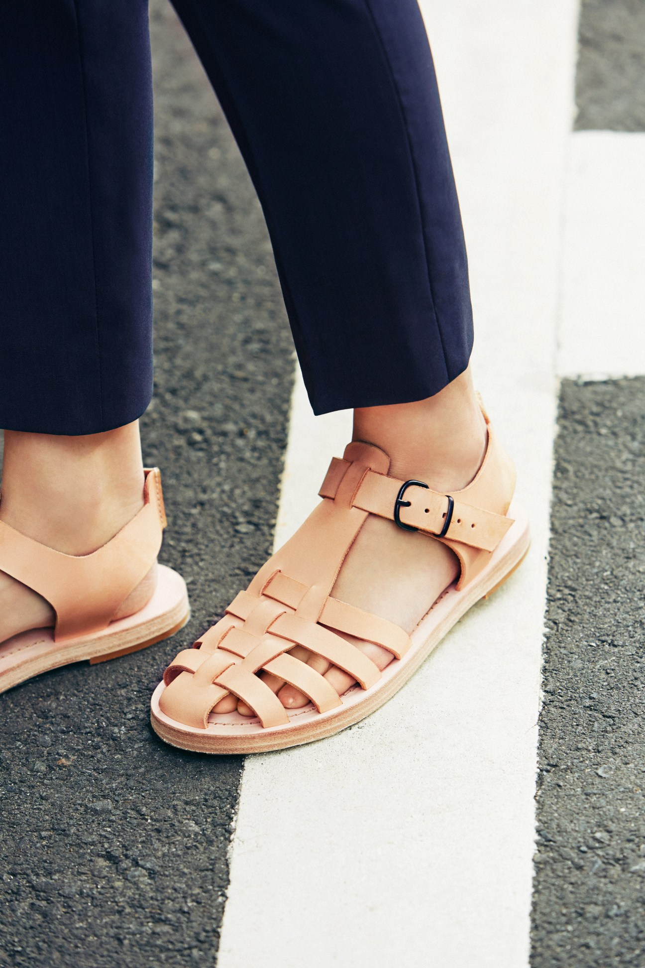 Person wearing navy trousers and tan leather strappy sandals standing on pavement.