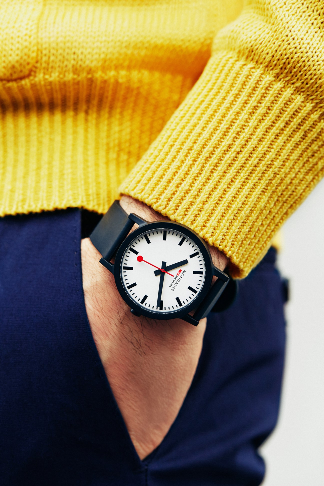 Man wearing yellow knit sweater and navy trousers with Mondaine Essence watch on wrist.