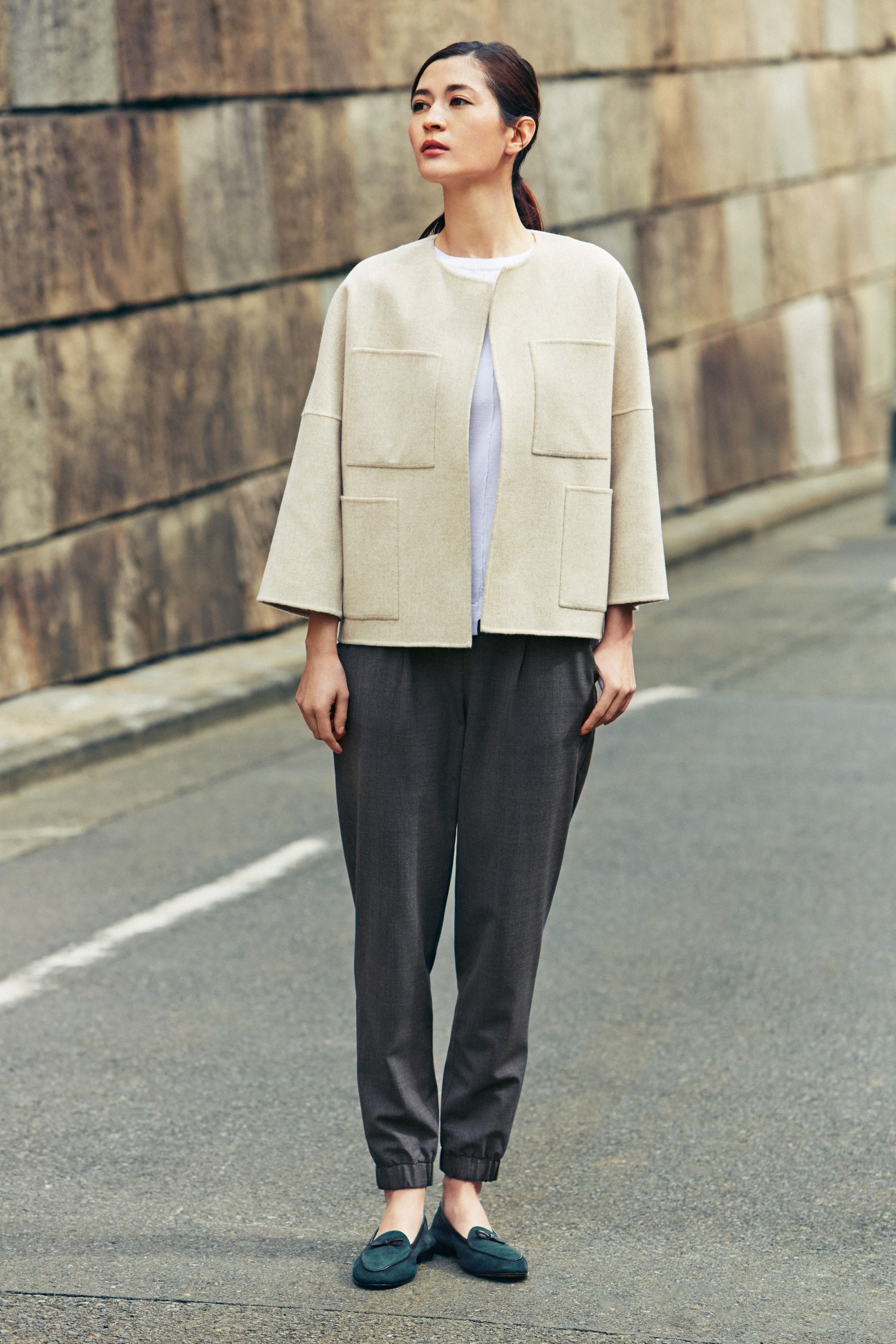 Woman wearing beige blazer with patch pockets, white t-shirt, black trousers, and dark loafers against brick wall.