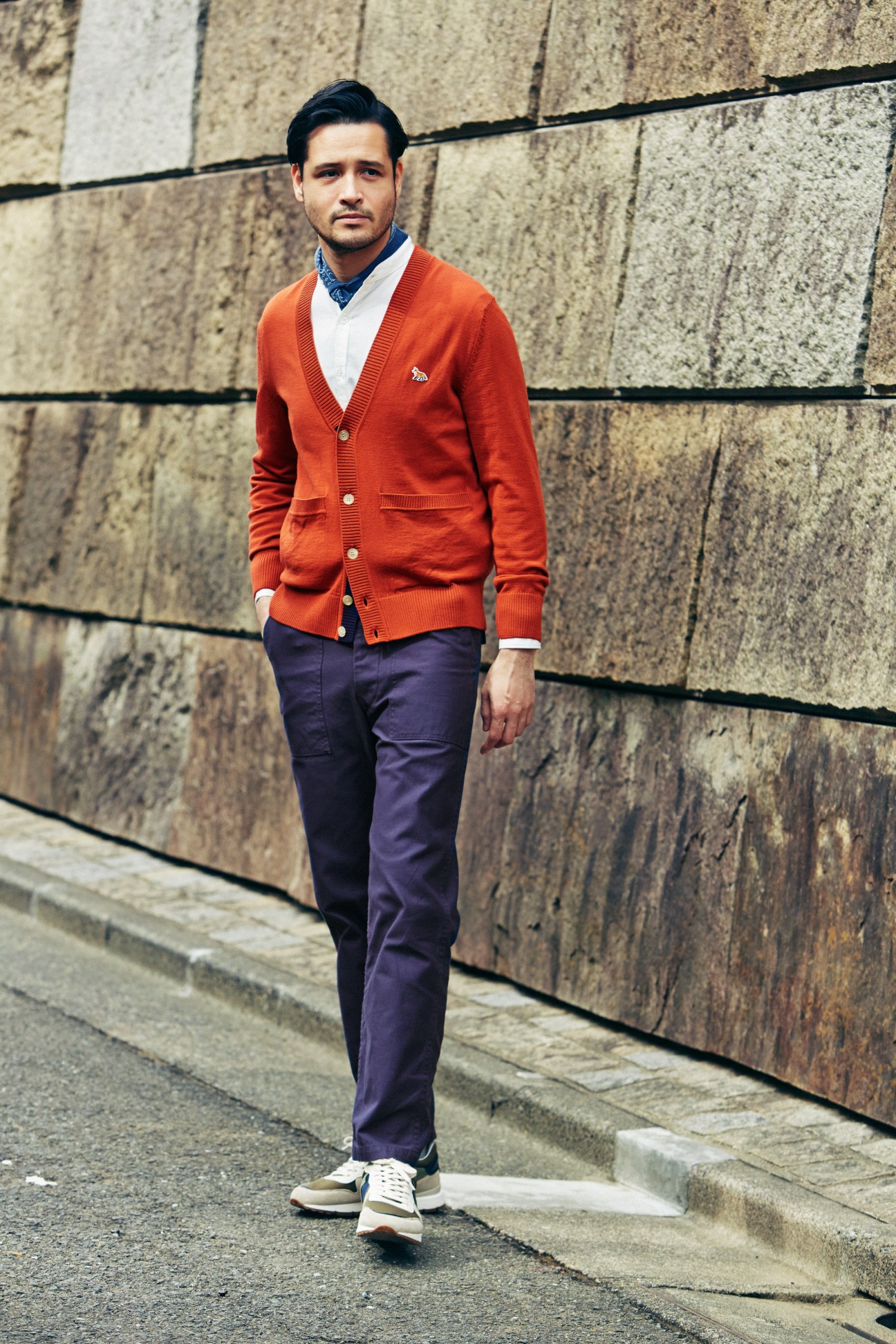 Man wearing a Maison Kitsuné cardigan, United Arrows shirt, Woolrich trousers and trainers, and Altea scarf against a stone wall.