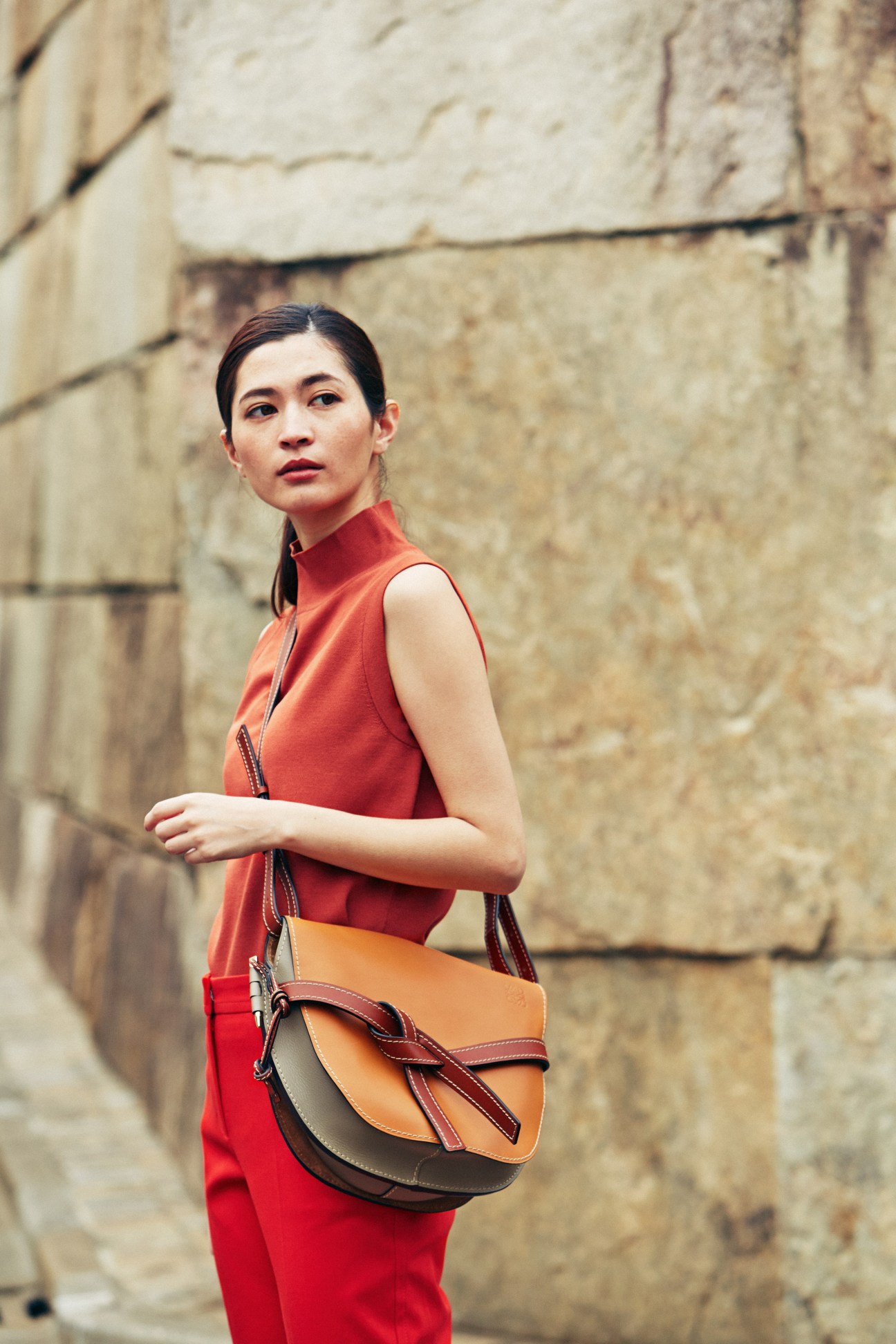 Woman in red sleeveless top and tan leather shoulder bag standing against stone wall.