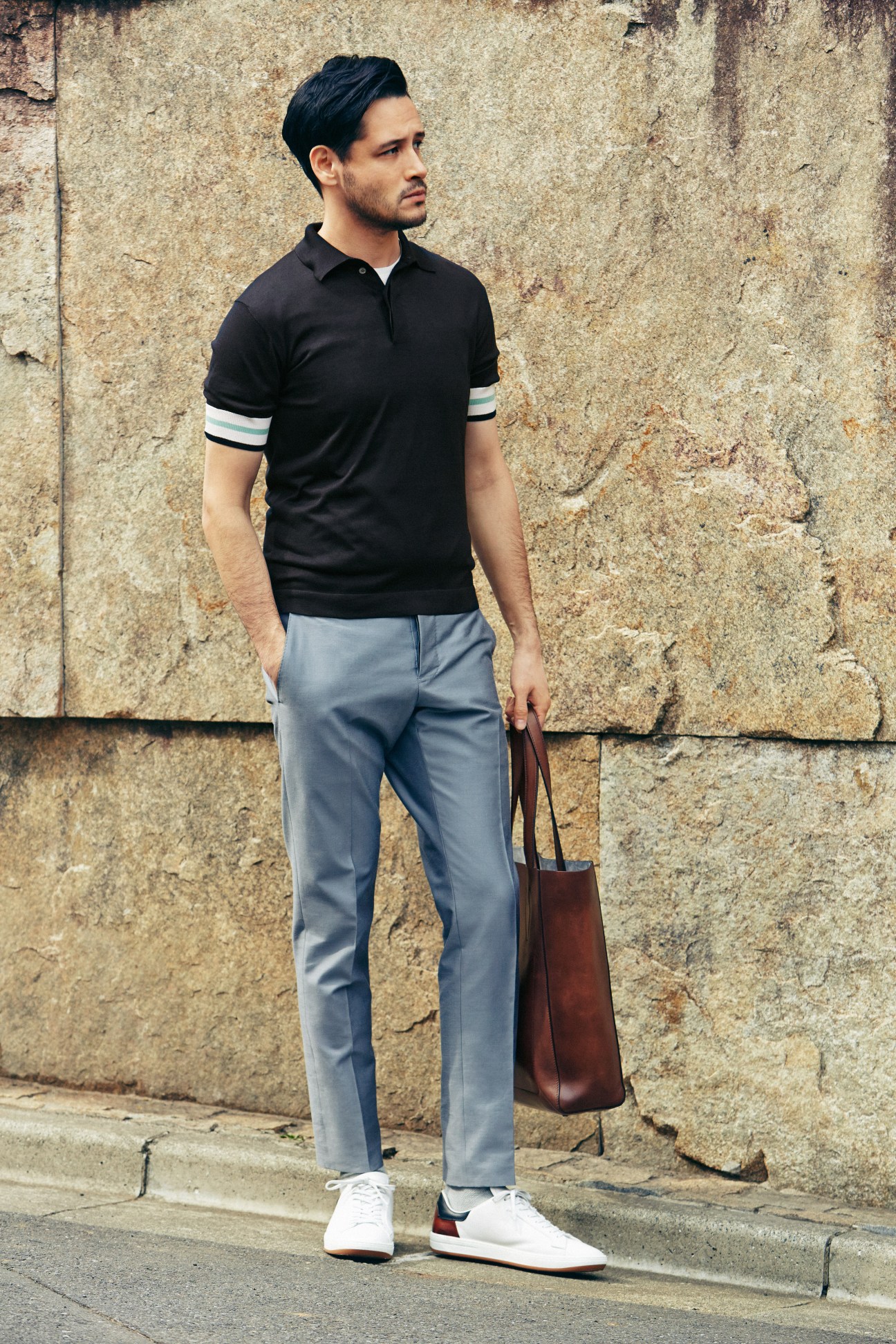 Man wearing black polo shirt with striped cuffs, light blue trousers, brown leather bag, and white sneakers against textured stone wall.
