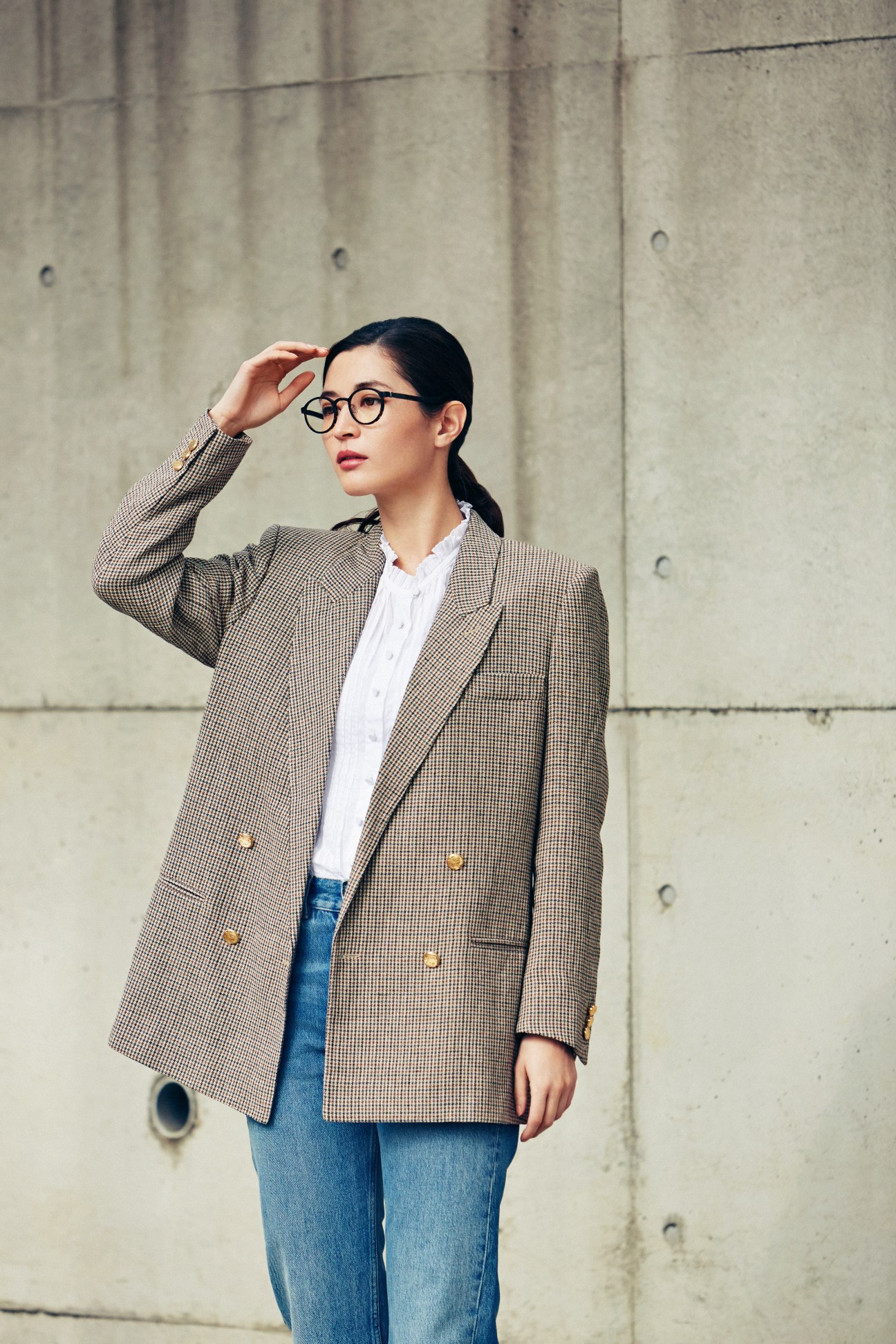 Woman wearing Gucci jacket and shirt with Acne Studios jeans and Lindberg glasses against concrete wall.