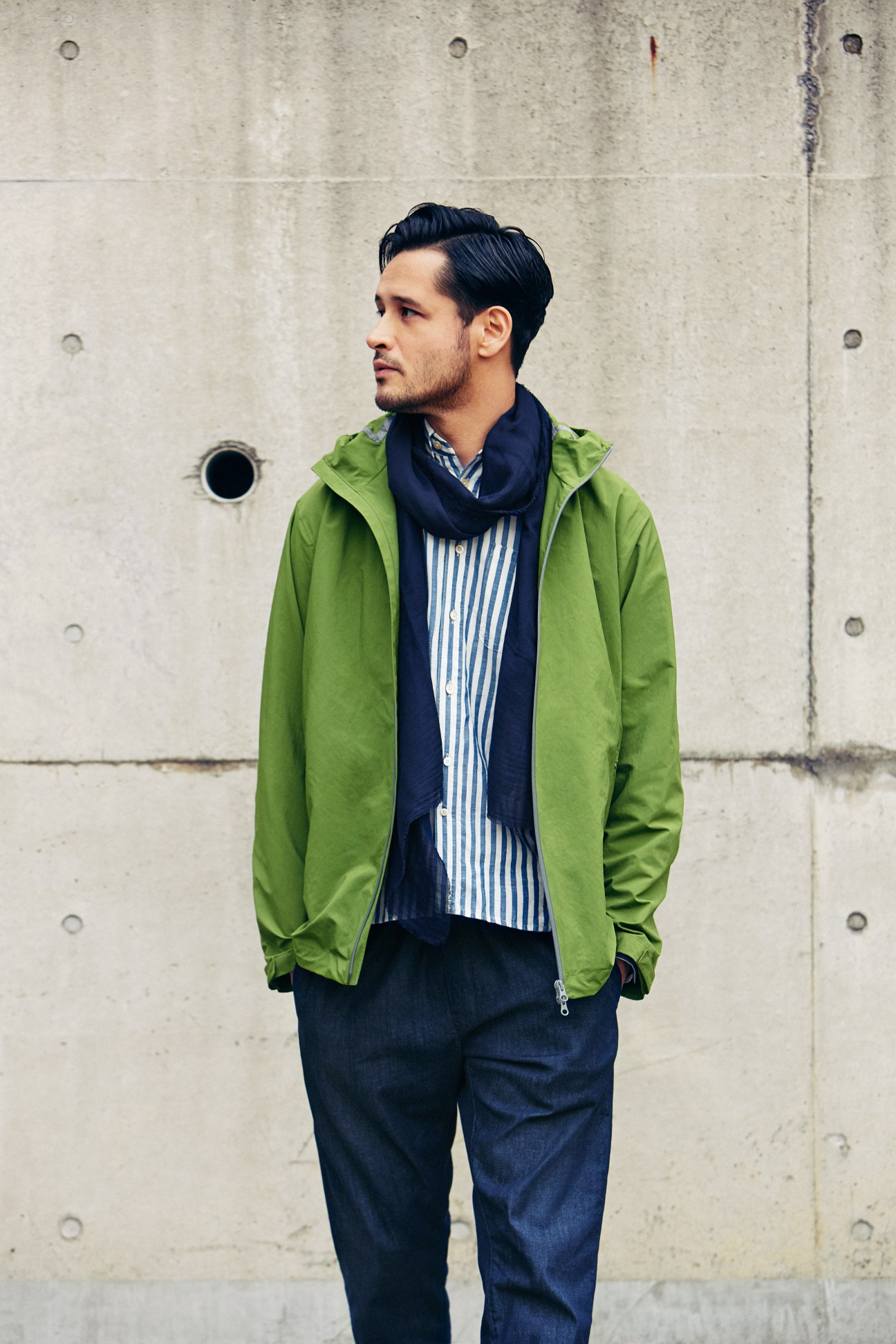 Man wearing a green jacket, navy striped shirt, and navy scarf against a concrete wall.