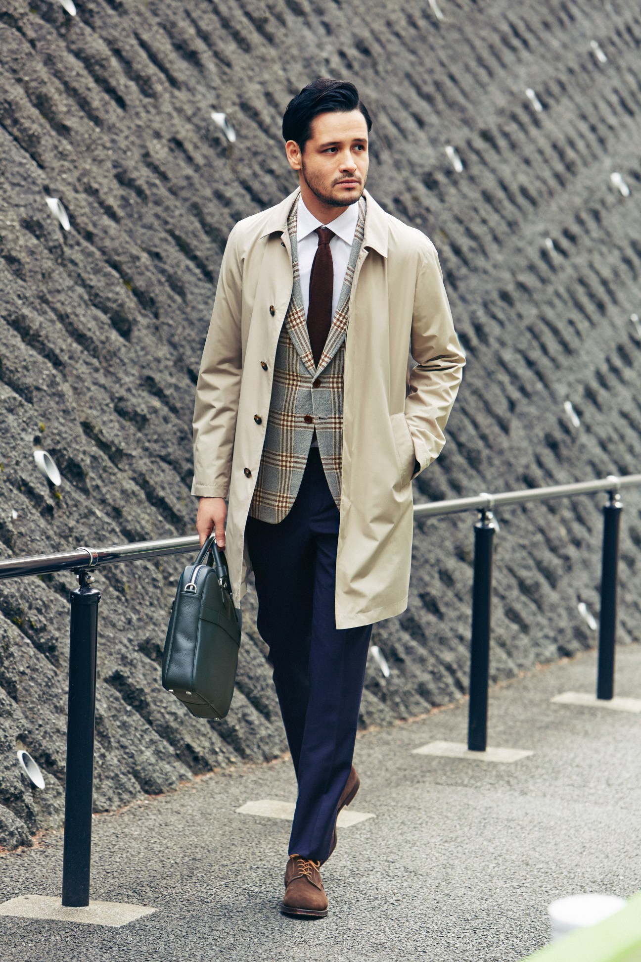 Man wearing beige coat, checked vest, dark tie, navy trousers, and brown shoes standing against textured brick wall.