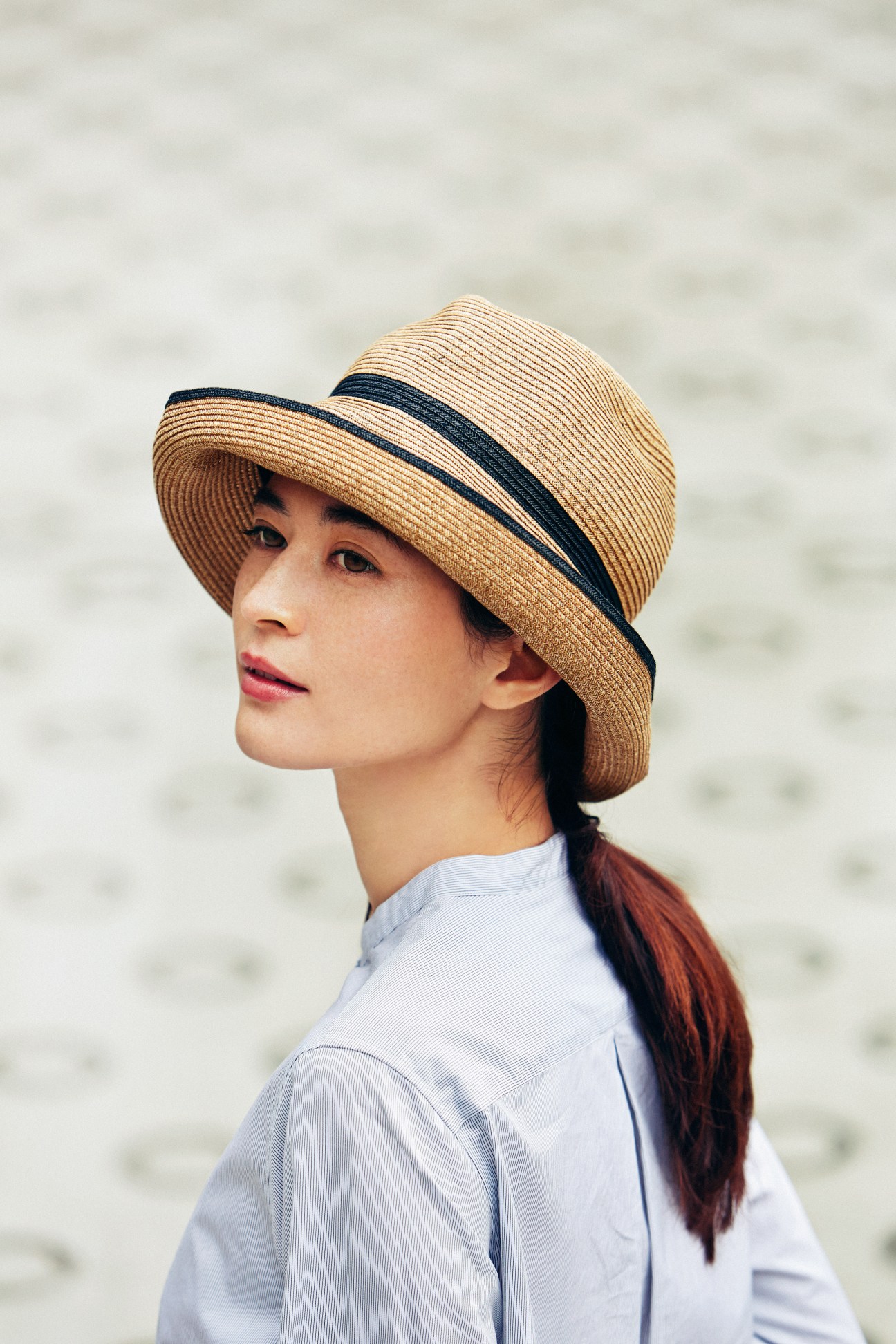 Woman wearing a straw hat with navy stripe and light blue shirt against neutral background.