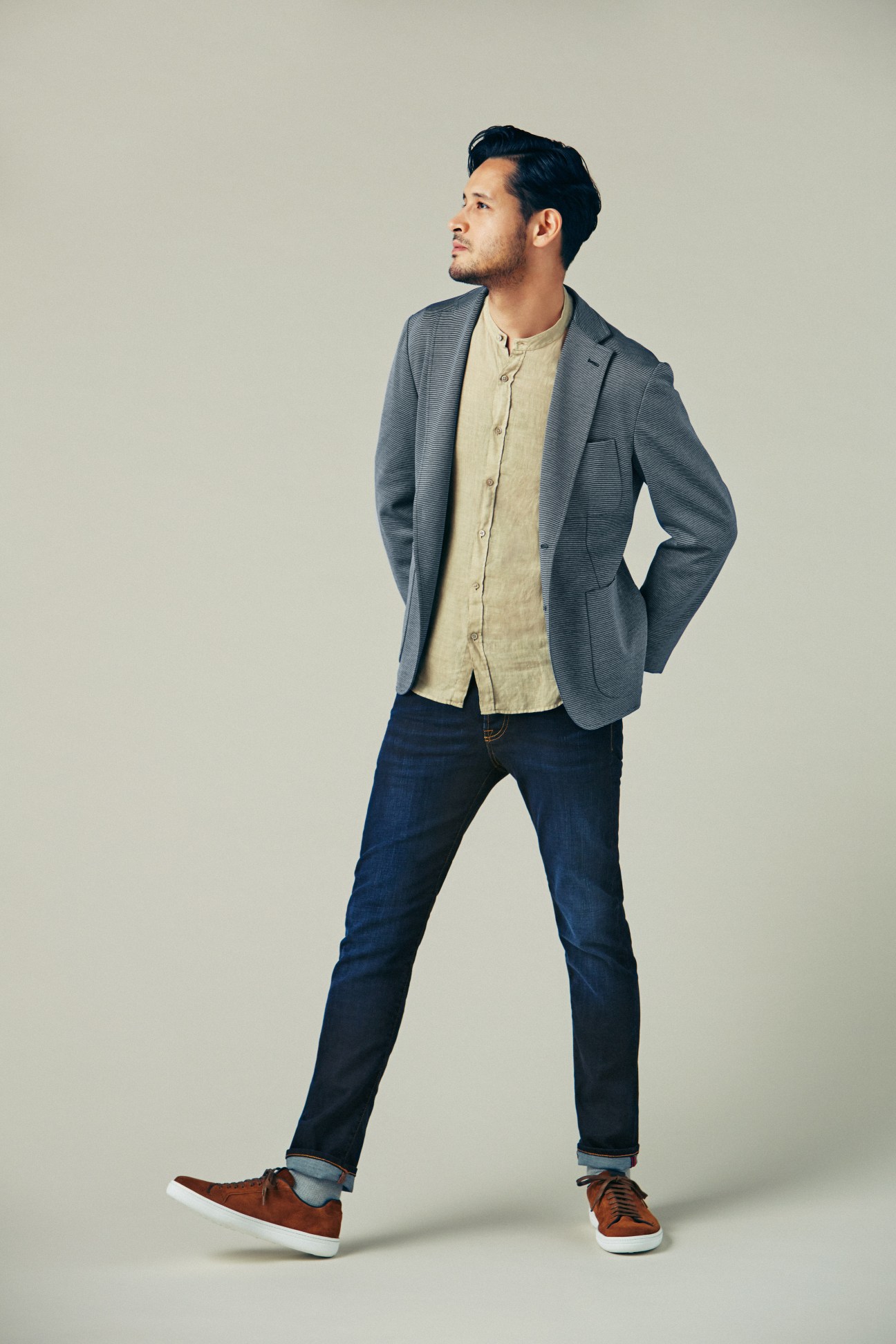 Man wearing gray blazer, cream button-up shirt, and rolled dark jeans with brown suede trainers.