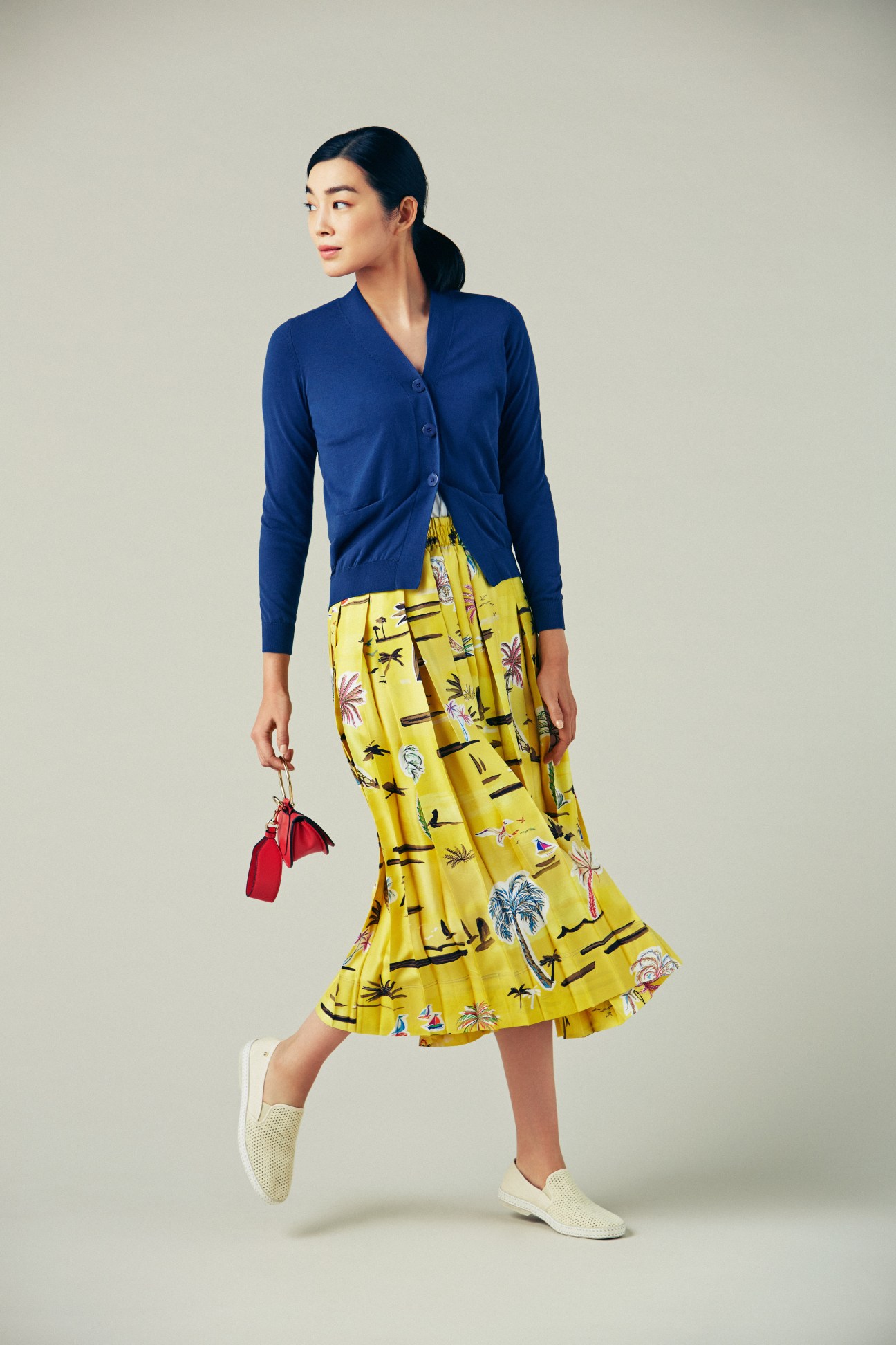 Woman wearing navy cardigan over yellow patterned skirt with white slip-on shoes and red handbag.