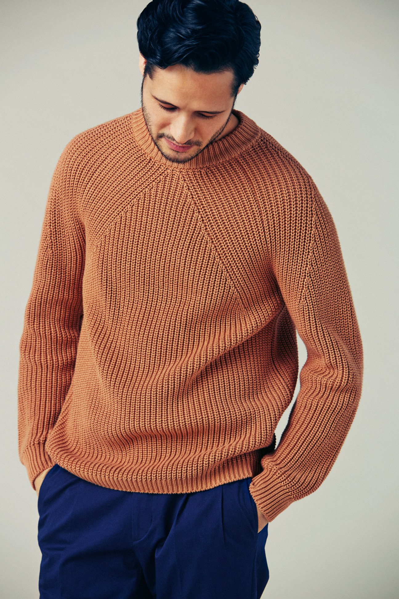 Man wearing an orange ribbed knit jumper and navy trousers, looking downward.