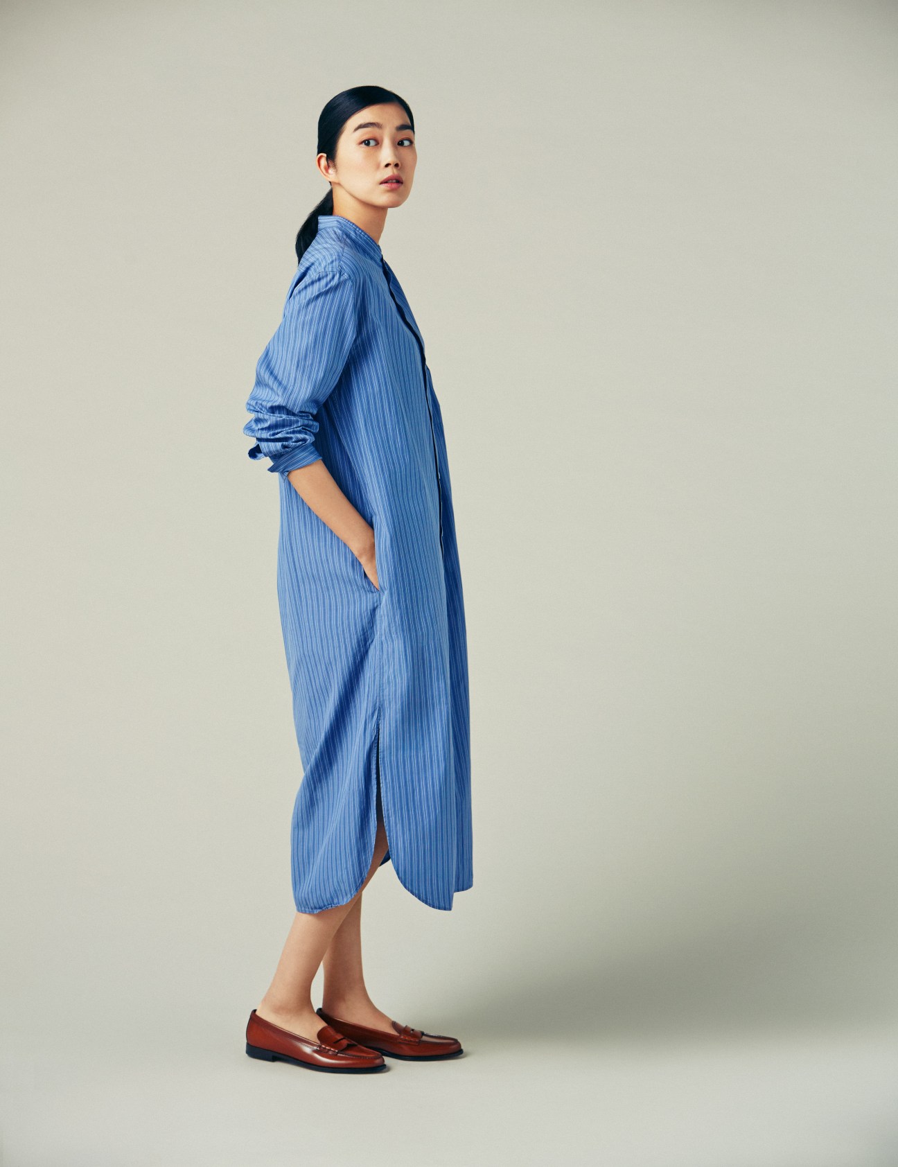 Woman wearing blue pinstriped coat dress with brown loafers, standing in profile against neutral background.