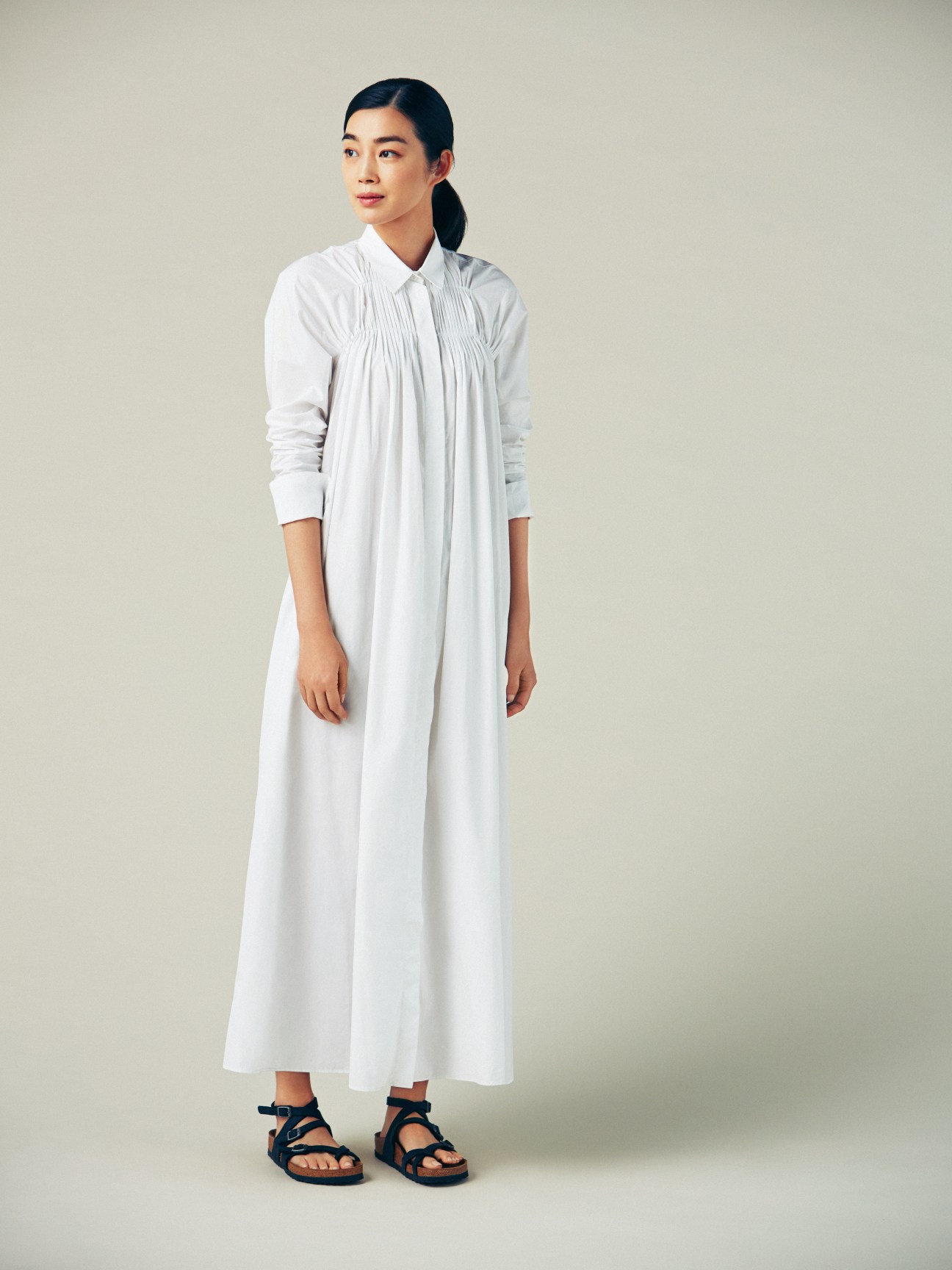 Woman in white pleated shirt dress and black sandals standing against neutral background.