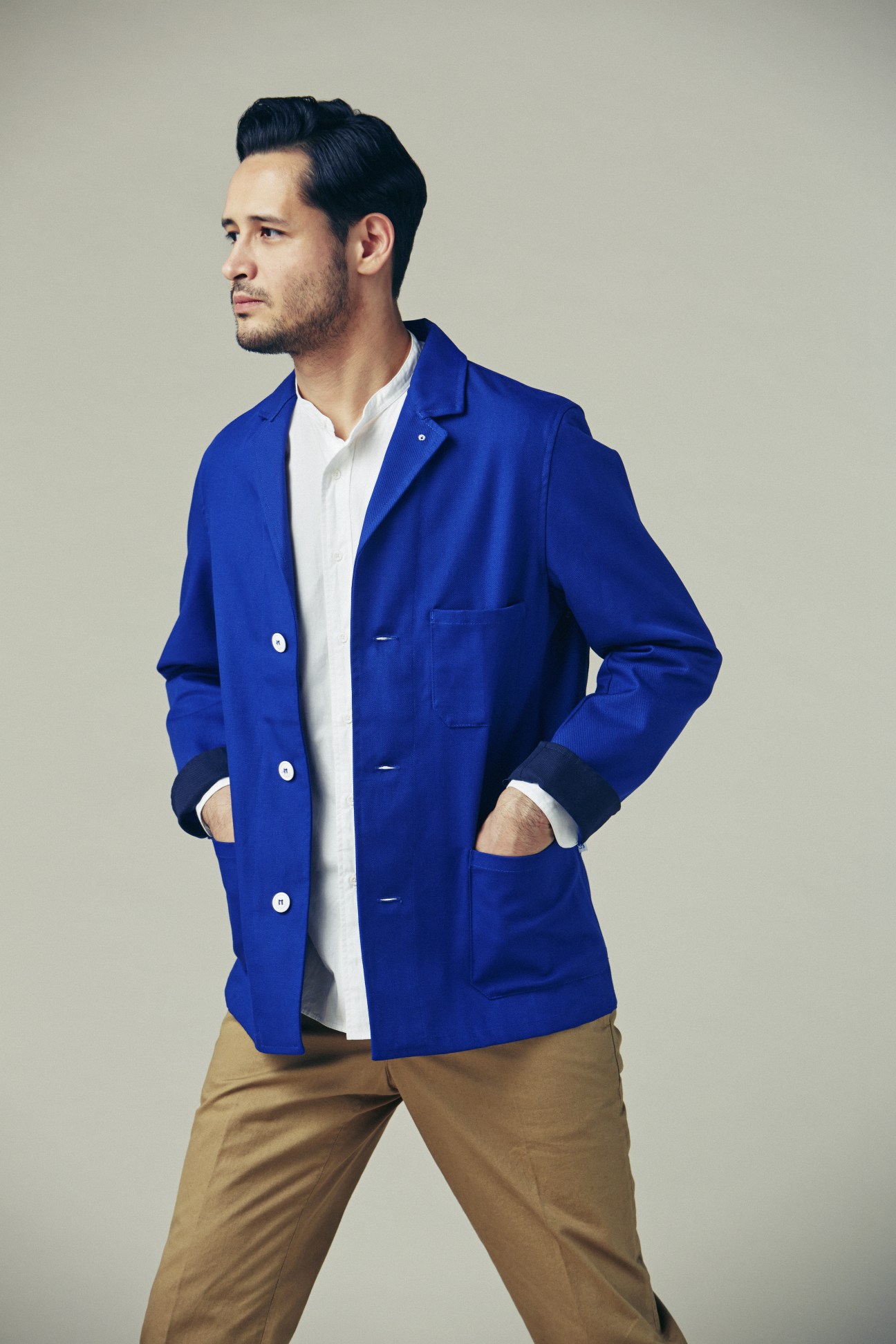 Man wearing royal blue blazer with white shirt and tan trousers, hands in pockets, posed against neutral background.