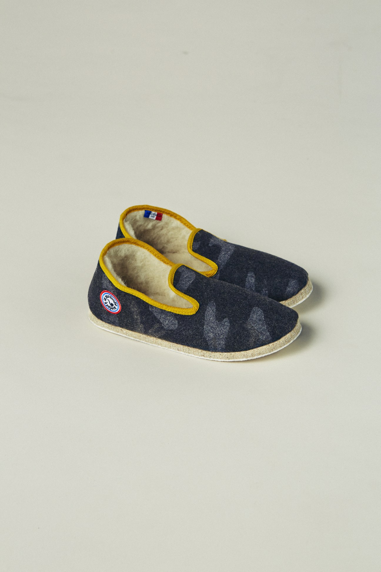 La Pantoufle à Pépère camo slippers with yellow trim and cream wool lining on neutral background.