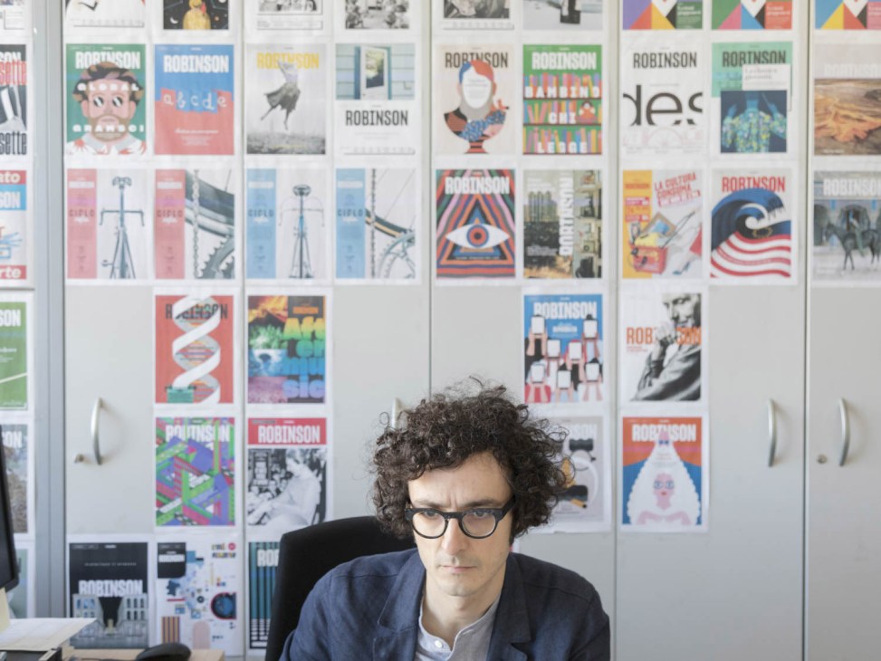 Man with curly dark hair and glasses sitting at desk in front of wall displaying magazine covers and design layouts.