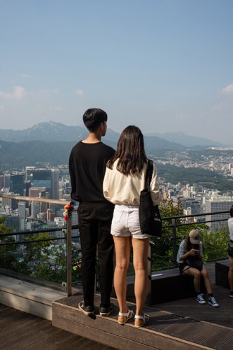 Two people stand at a viewpoint overlooking Seoul's skyline and mountains at sunrise.