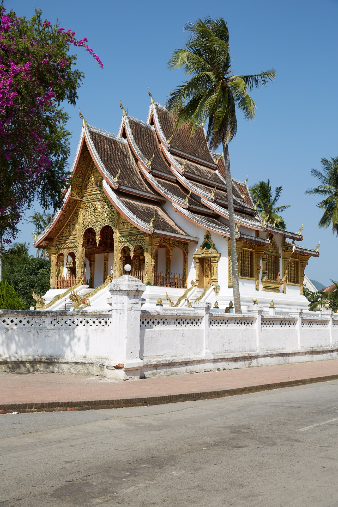 Ornate Buddhist temple with gilded details, white walls, tiered roofs and palm trees in Luang Prabang