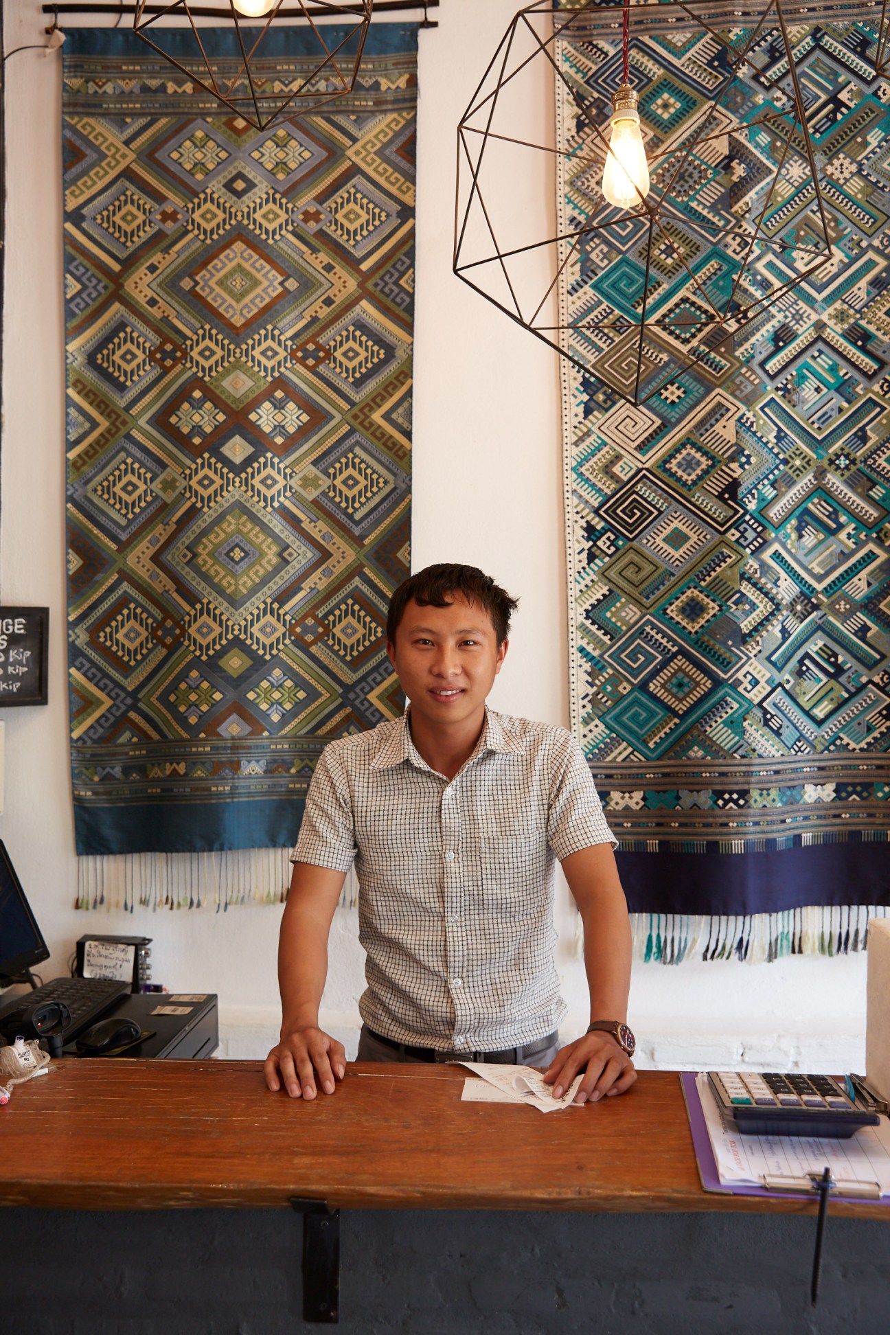 Man standing at wooden counter in front of traditional woven Laotian textiles displayed on white wall.
