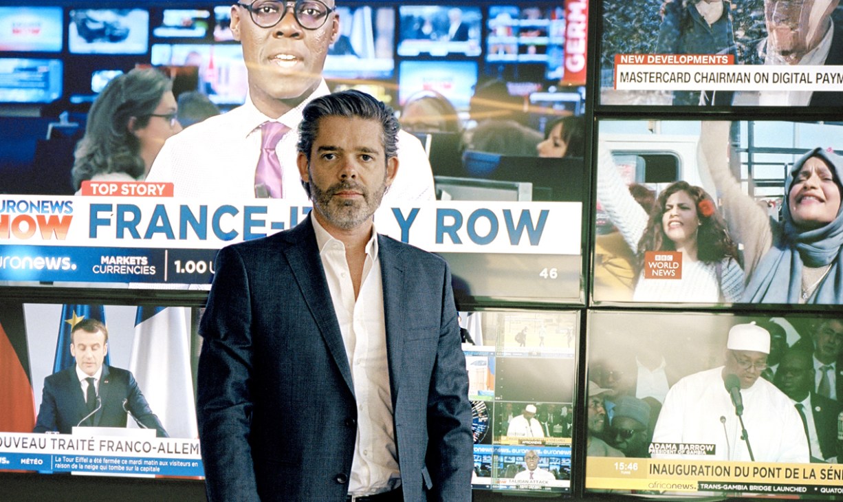 Michael Peters, Euronews CEO, stands in front of multiple broadcast monitors displaying news content in the Lyon newsroom.
