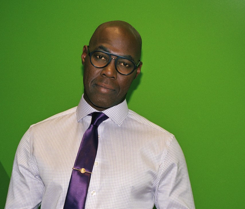 Tokunbo Salako, Euronews news anchor, wearing glasses and a purple tie against a green background.
