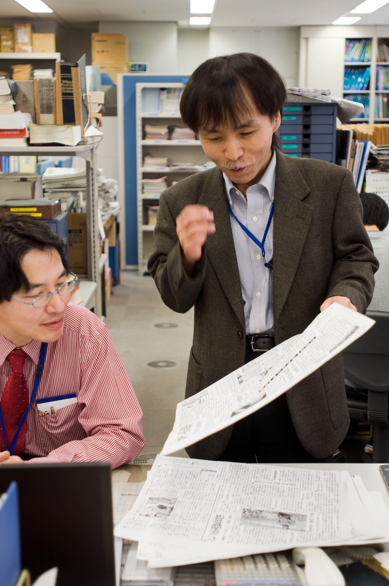 Two Japanese newspaper staff members review printed pages in an office with shelving and filing cabinets.