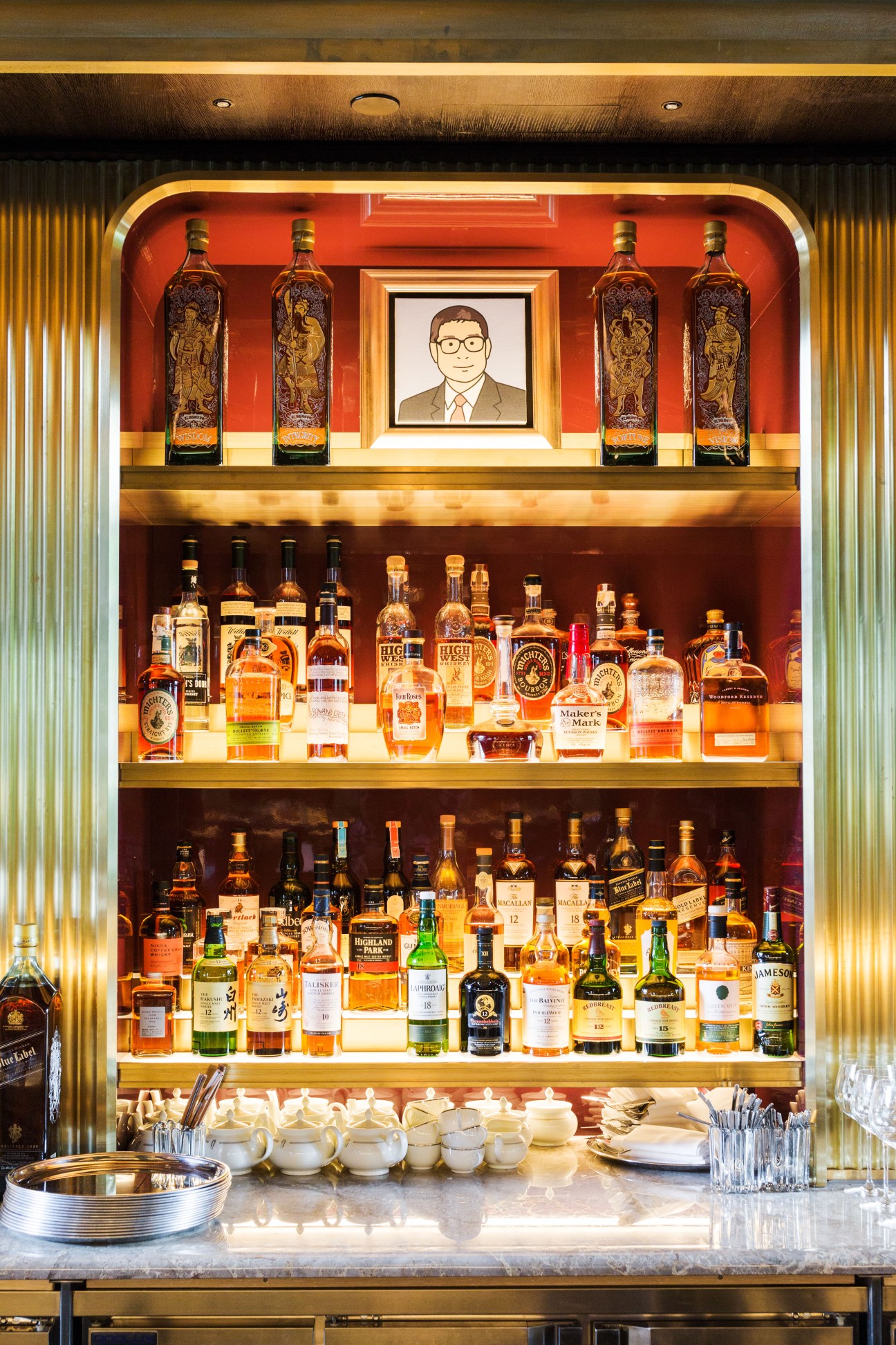 Illuminated bar display with whiskey bottles and portrait illustration at Atlas Bar in Singapore.