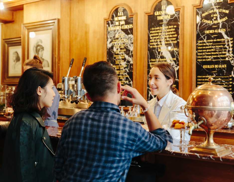 Staff member serves customers at Grand Café Tortoni counter in wood-paneled interior with black menus.