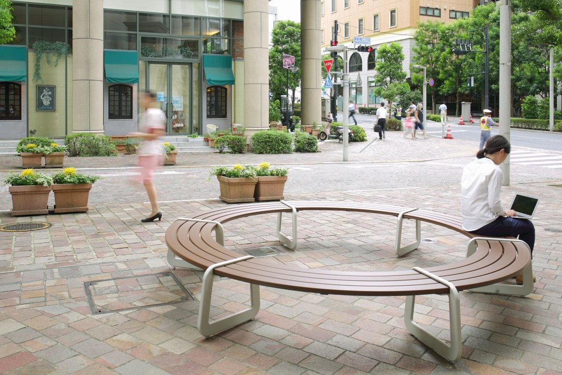 Modern curved wooden bench with metal supports in an urban plaza with pedestrians and greenery.
