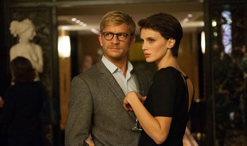 A man in glasses and grey jacket stands with a woman in black shirt in an indoor setting.