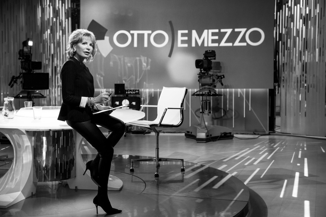 Lilli Gruber on the set of her Italian talk show Otto e Mezzo, seated at a desk with studio equipment visible behind her.
