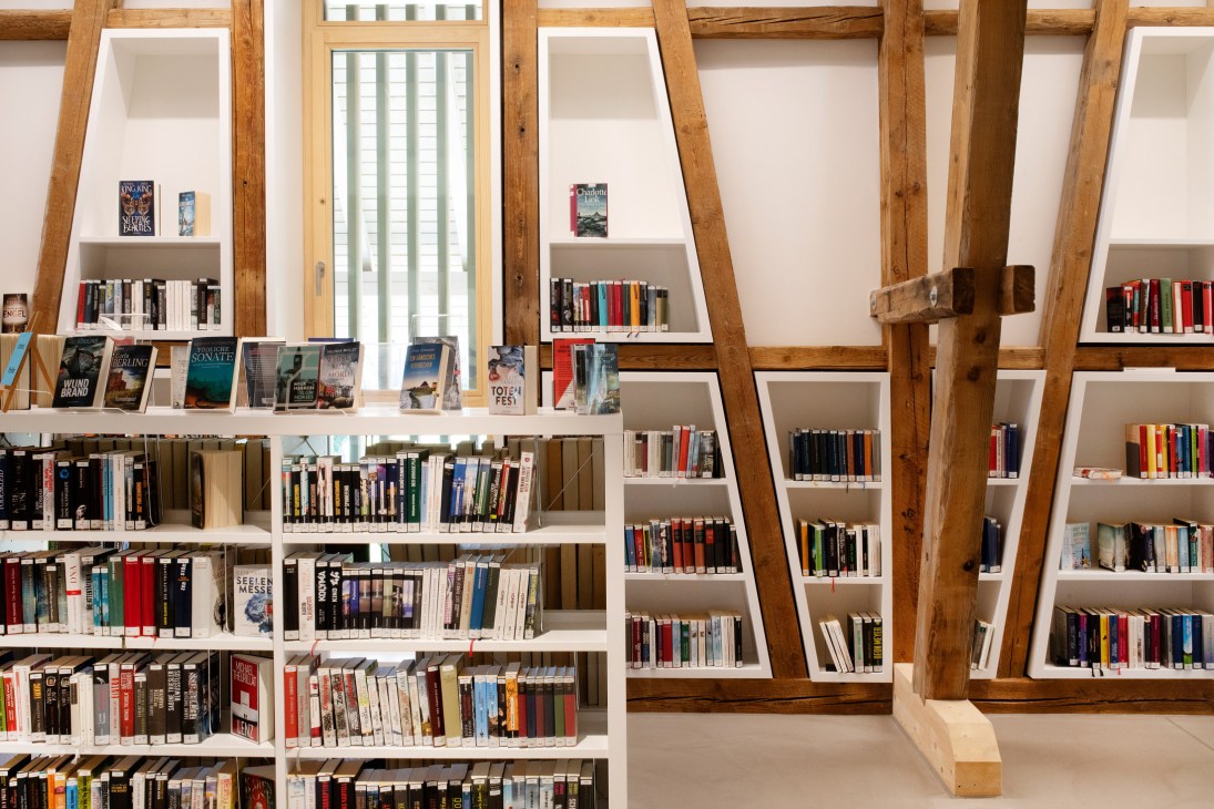 Interior of Bibliothek Kressbronn library with wooden beams, white shelving units filled with colorful books, and natural light from windows.