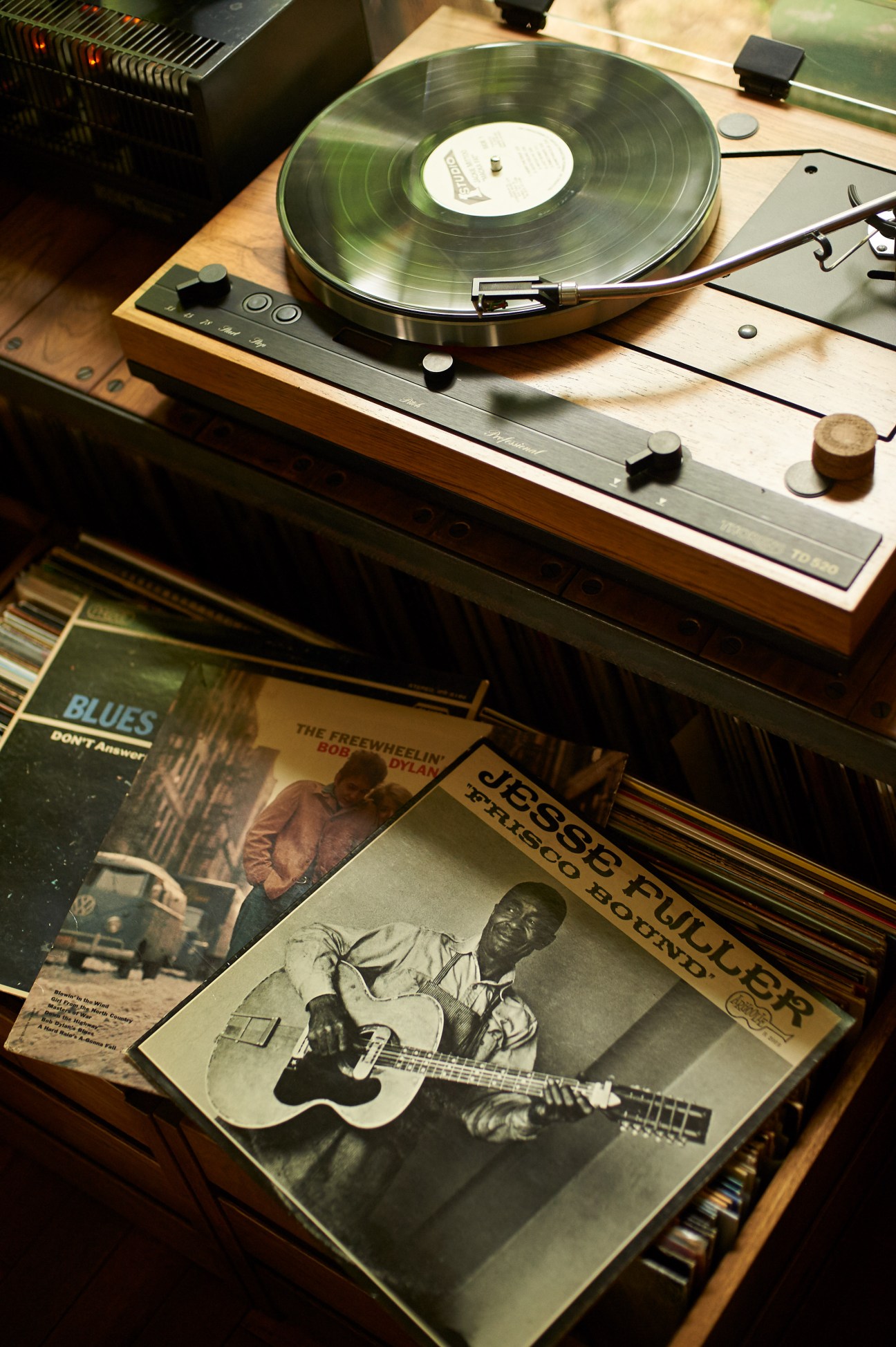 A Thorens turntable playing a vinyl record with blues albums displayed below on wooden furniture.