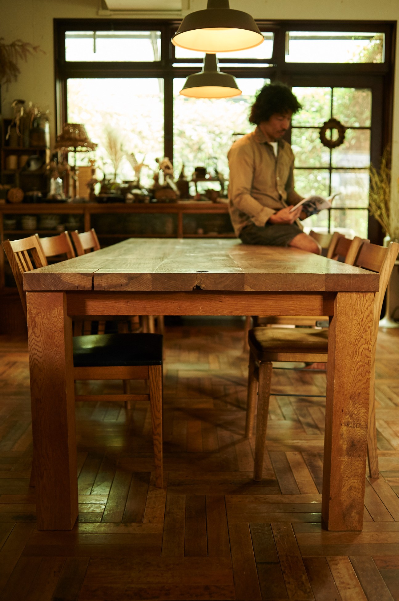 Tokuhiko Kise sits at a wooden dining table with natural cracks and knots in a bright room with large windows.