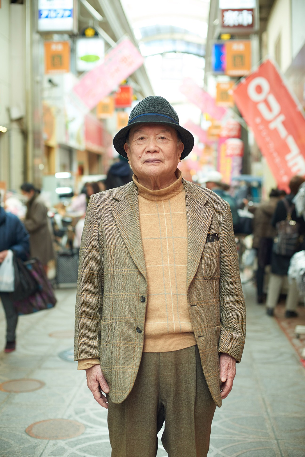 Elderly man in beige blazer and hat standing in Japanese shopping arcade