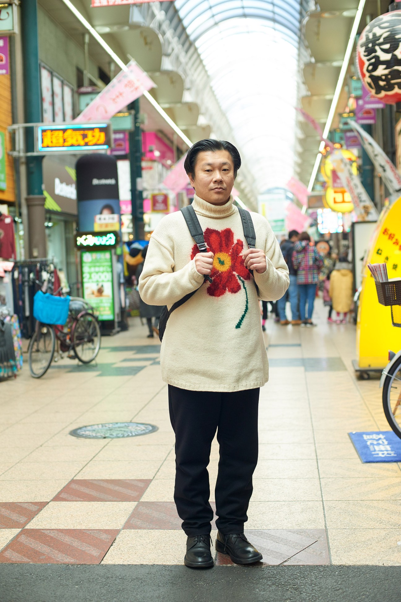 Man wearing cream sweater with red flower design standing in covered shopping arcade in Osaka.