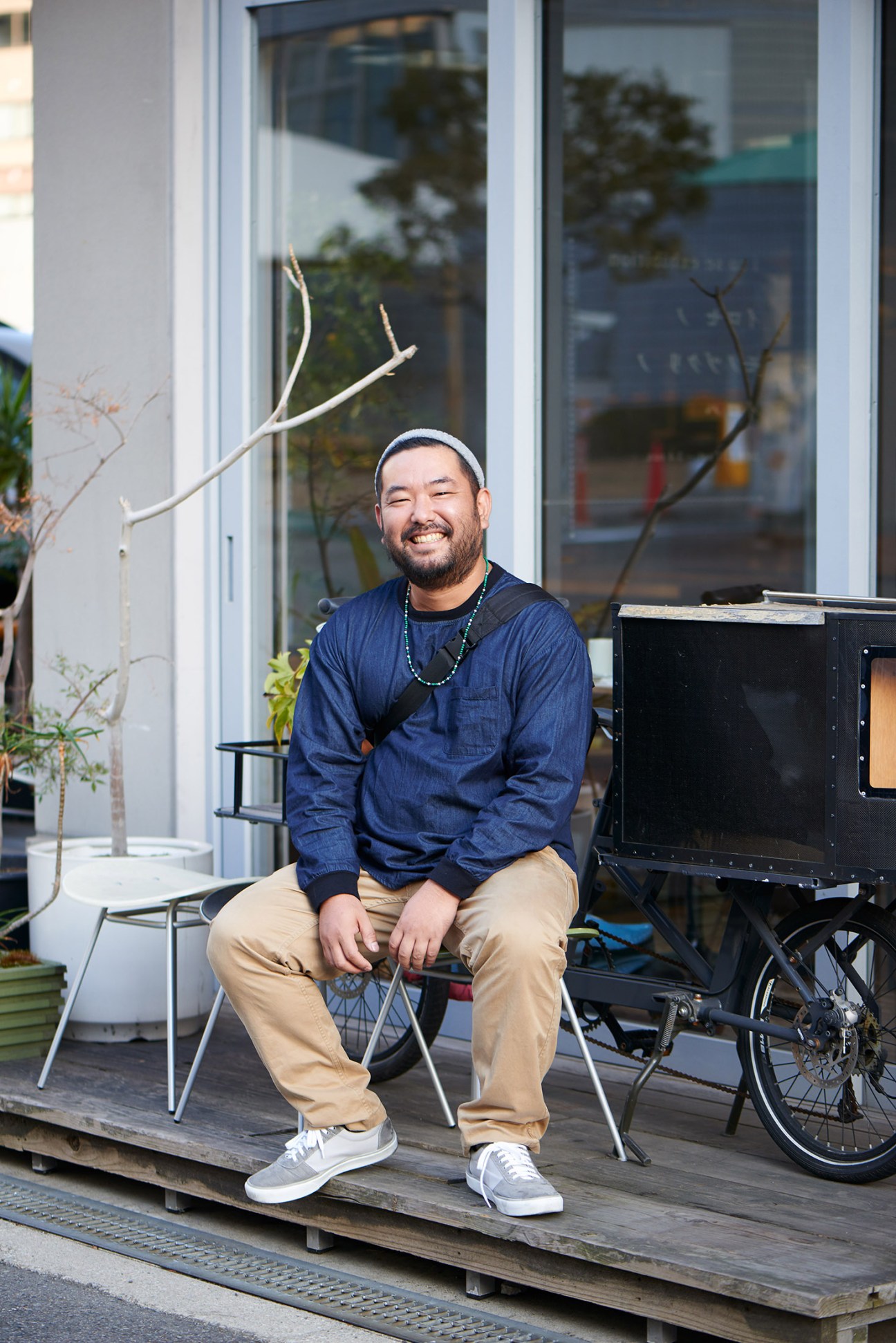 Man in blue denim shirt and white headband sitting on wooden platform outside storefront with bicycle and potted plants.