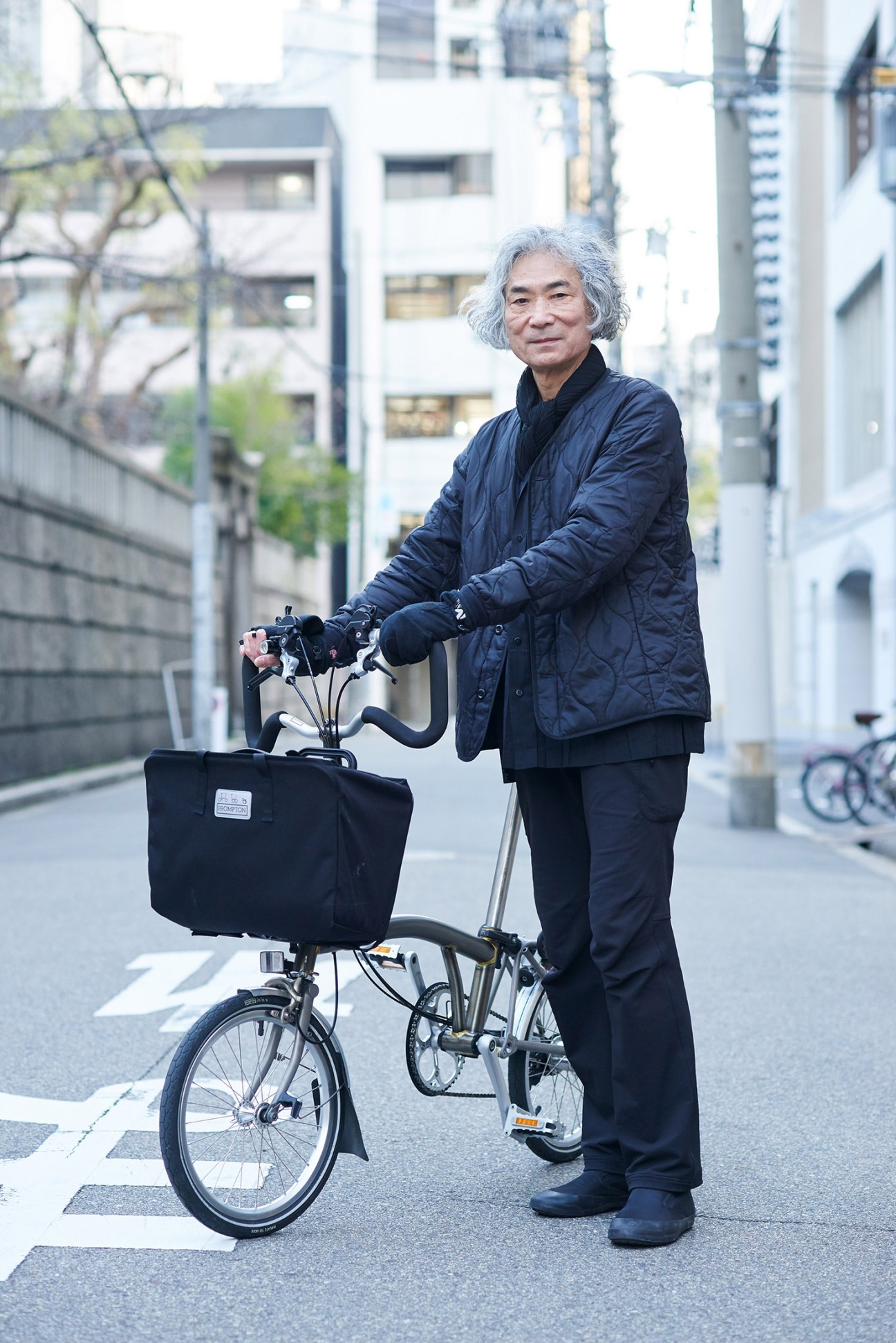 Elderly man with gray hair standing with a bicycle carrying a black cargo basket on a city street.