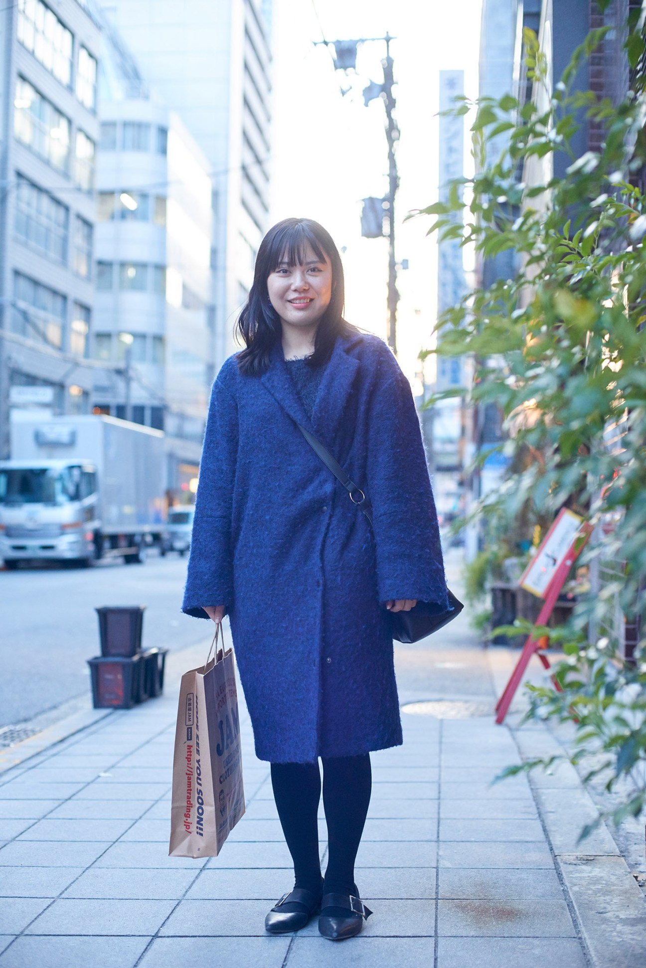 Young woman in blue coat holding shopping bag on Osaka street with buildings and greenery