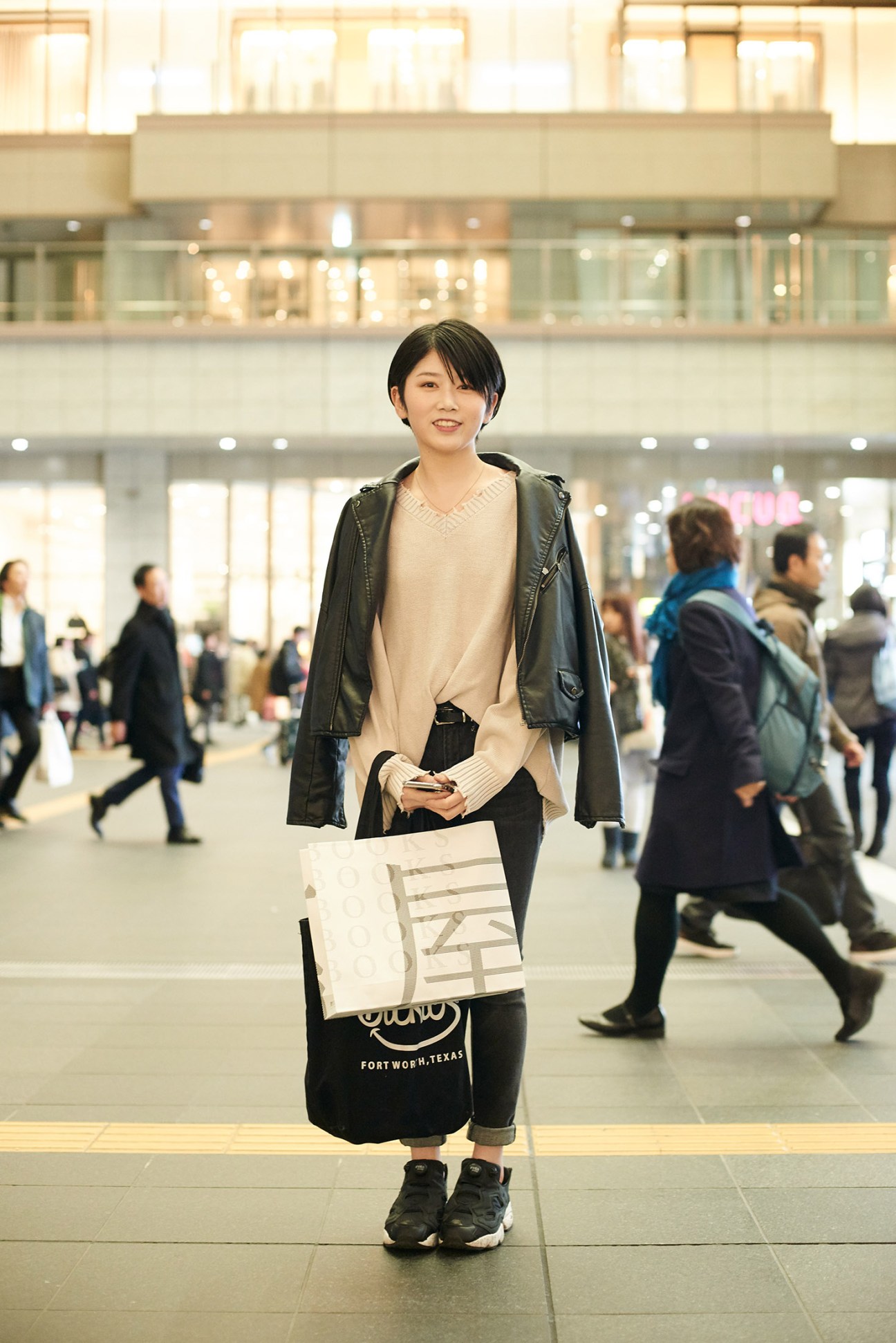 Young woman in cream sweater and dark jacket holding shopping bag in modern shopping mall