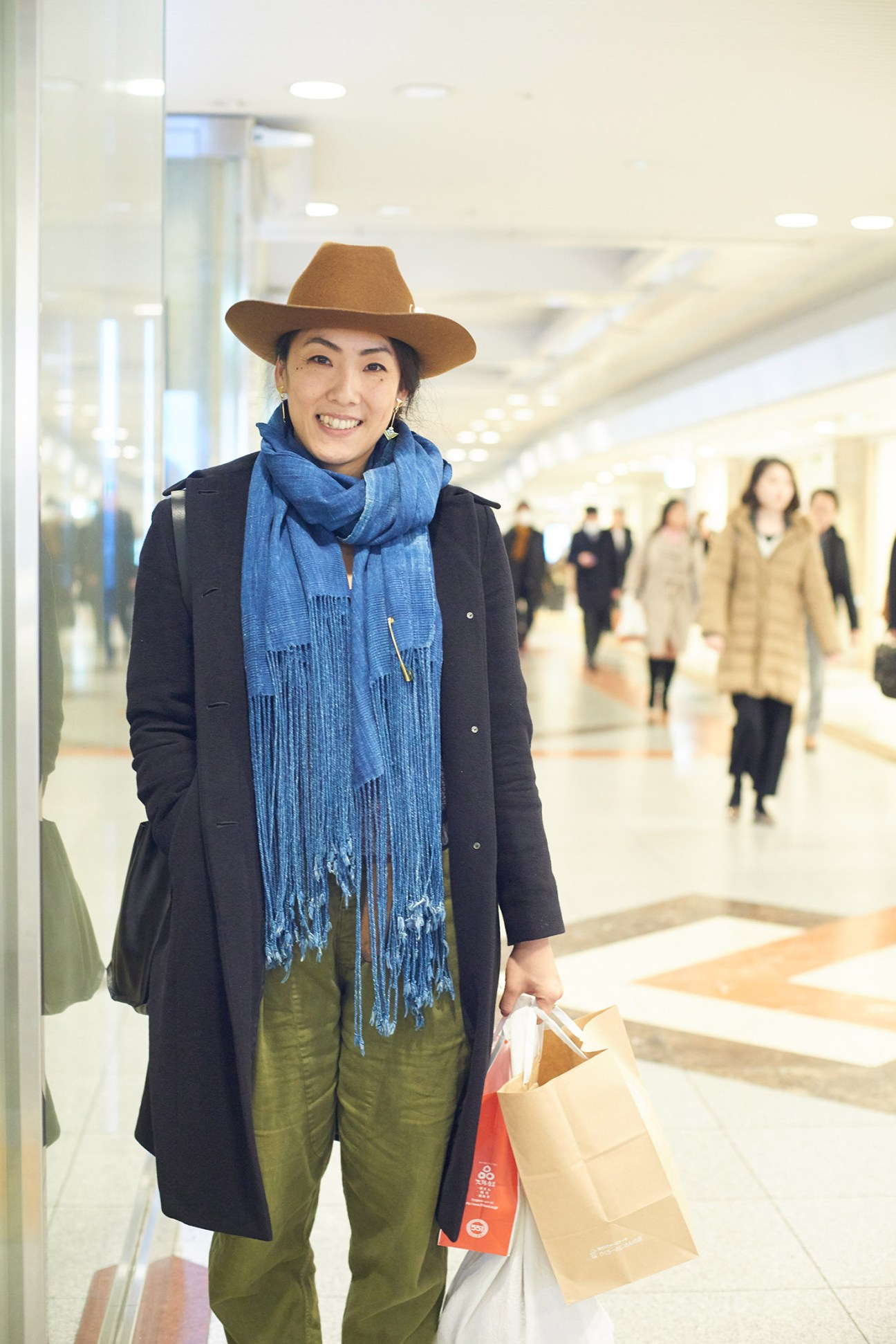 Woman wearing brown cowboy hat, blue scarf, black coat, and olive pants stands in modern shopping mall holding shopping bags.