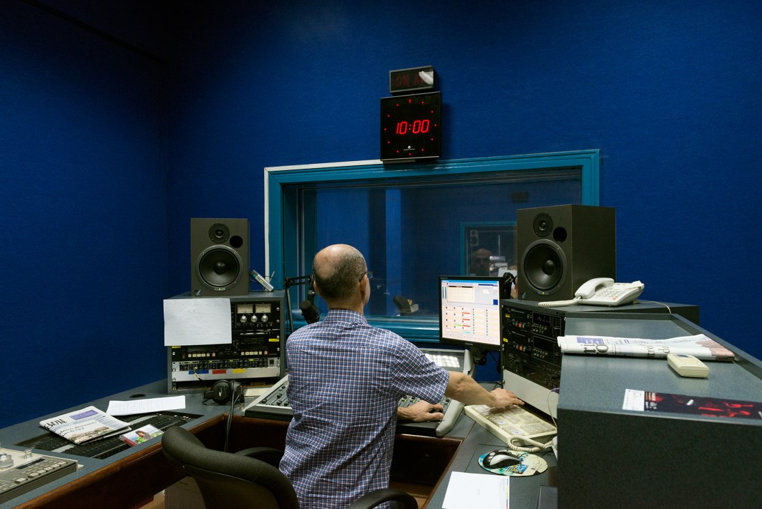 Radio technician working at control desk in broadcast studio with blue walls and monitoring equipment.