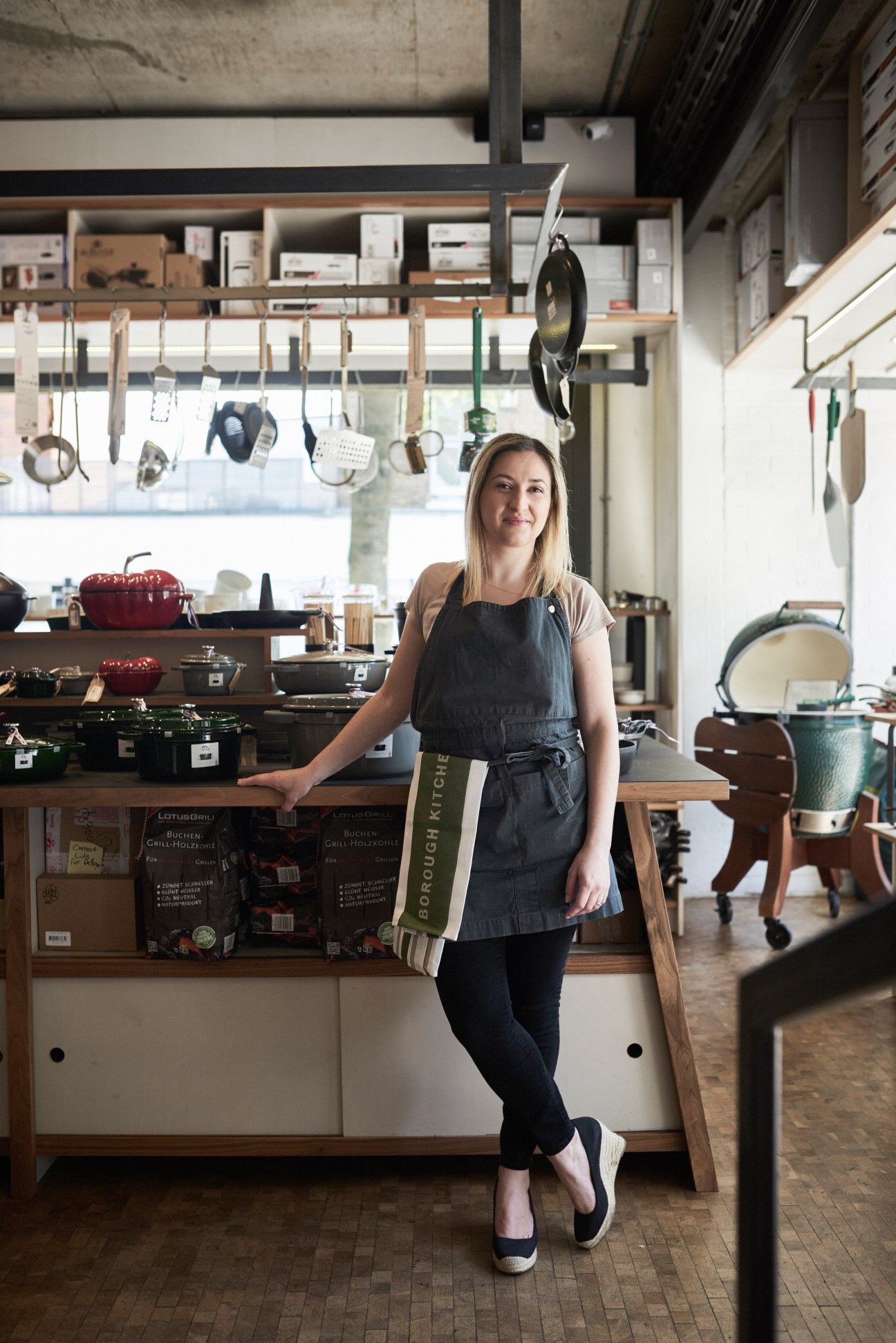 Woman in apron standing in modern kitchen showroom with hanging cookware and shelves of kitchen products