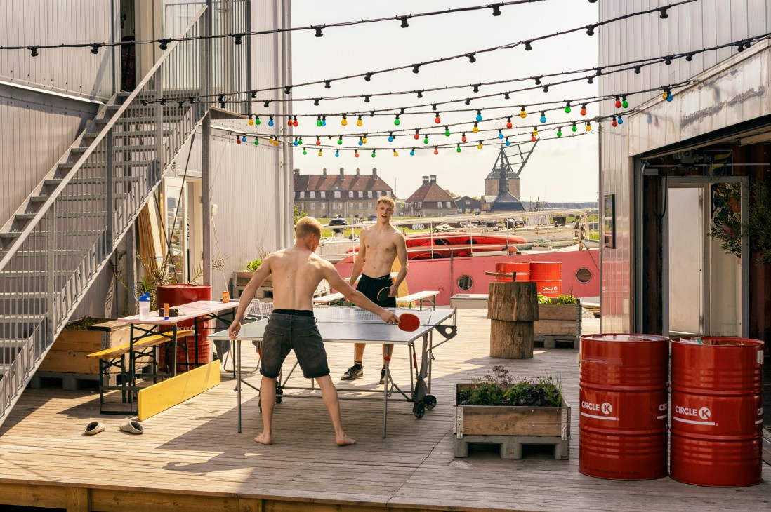 Two shirtless men play table tennis on a rooftop terrace with colorful string lights overhead in Copenhagen.