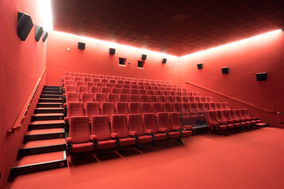 Modern cinema auditorium with red seats and walls, linear ceiling lights, and labeled seating rows.