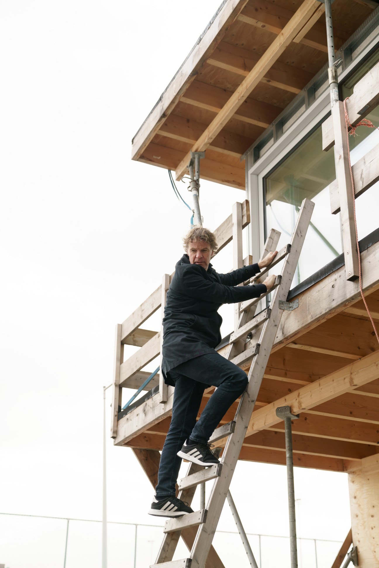 Man climbing a wooden ladder on a construction site with exposed timber framing overhead.