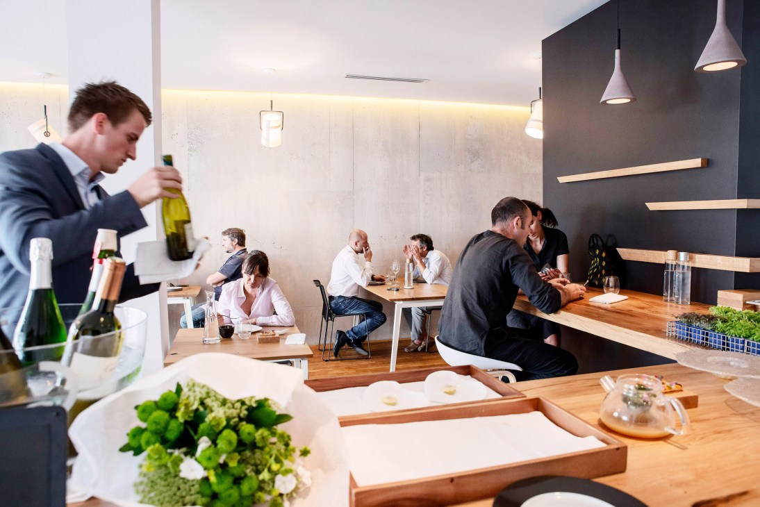 Interior of a modern restaurant with diners at tables, a server holding wine, and minimalist décor featuring wooden tables and pendant lights.