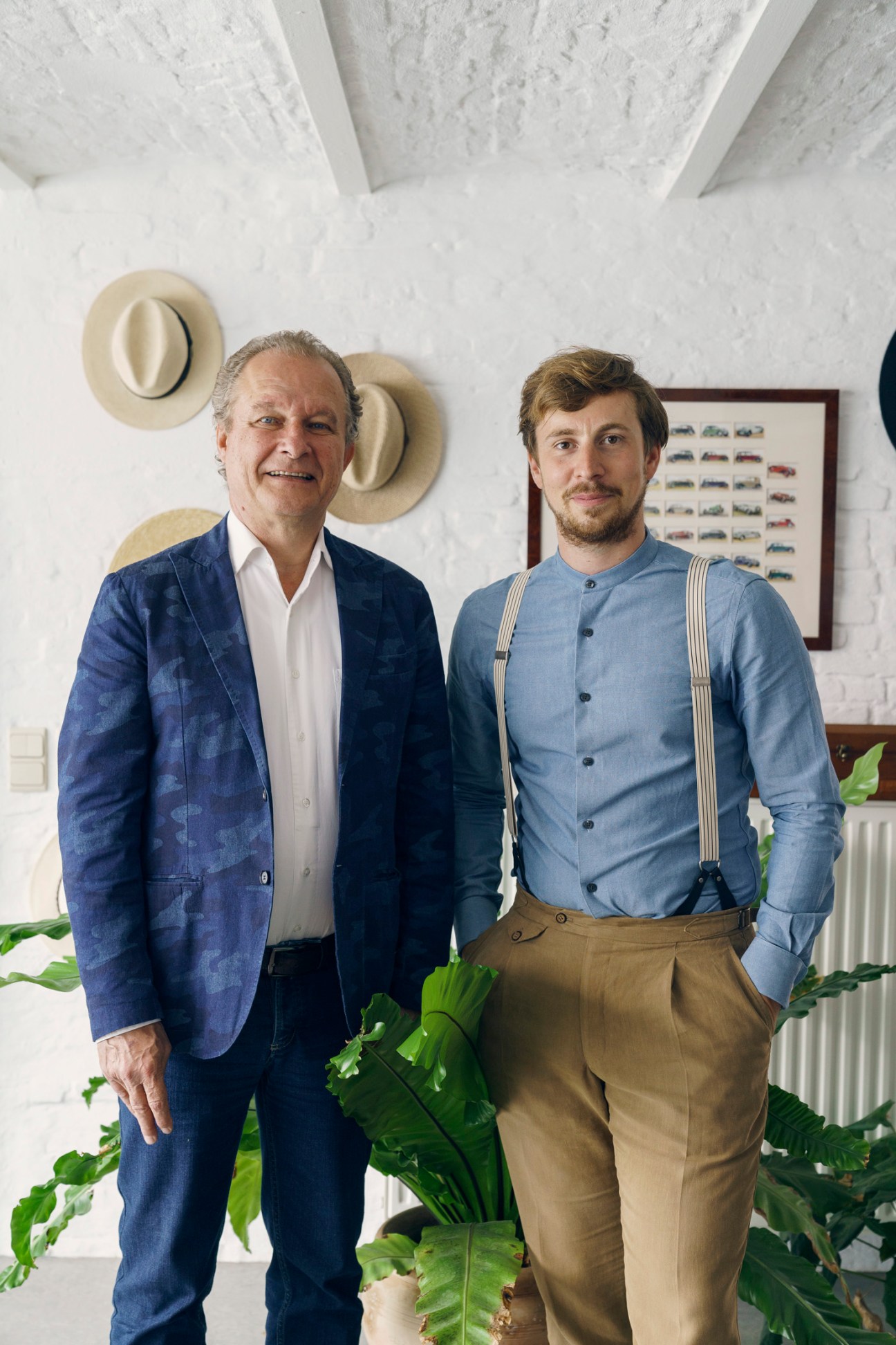 Two men posing in a white-walled interior space with hats and artwork behind them.