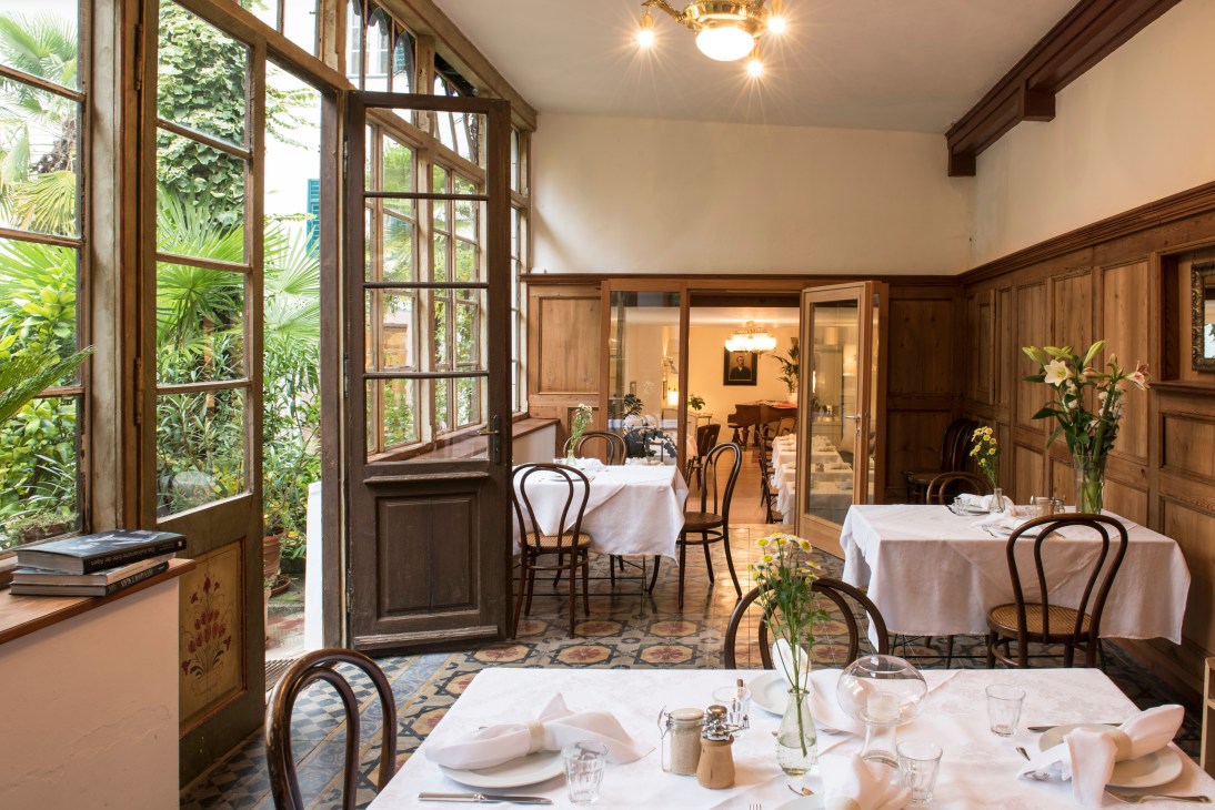 Fine dining restaurant interior with wooden paneling, white tablecloths, bentwood chairs, fresh flowers, and garden views through open doors.