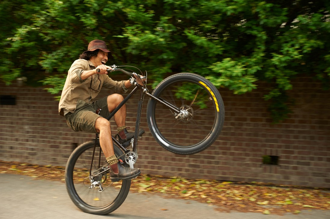 Man riding a bicycle with one wheel lifted off the ground in a playful wheelie near a brick wall and trees.